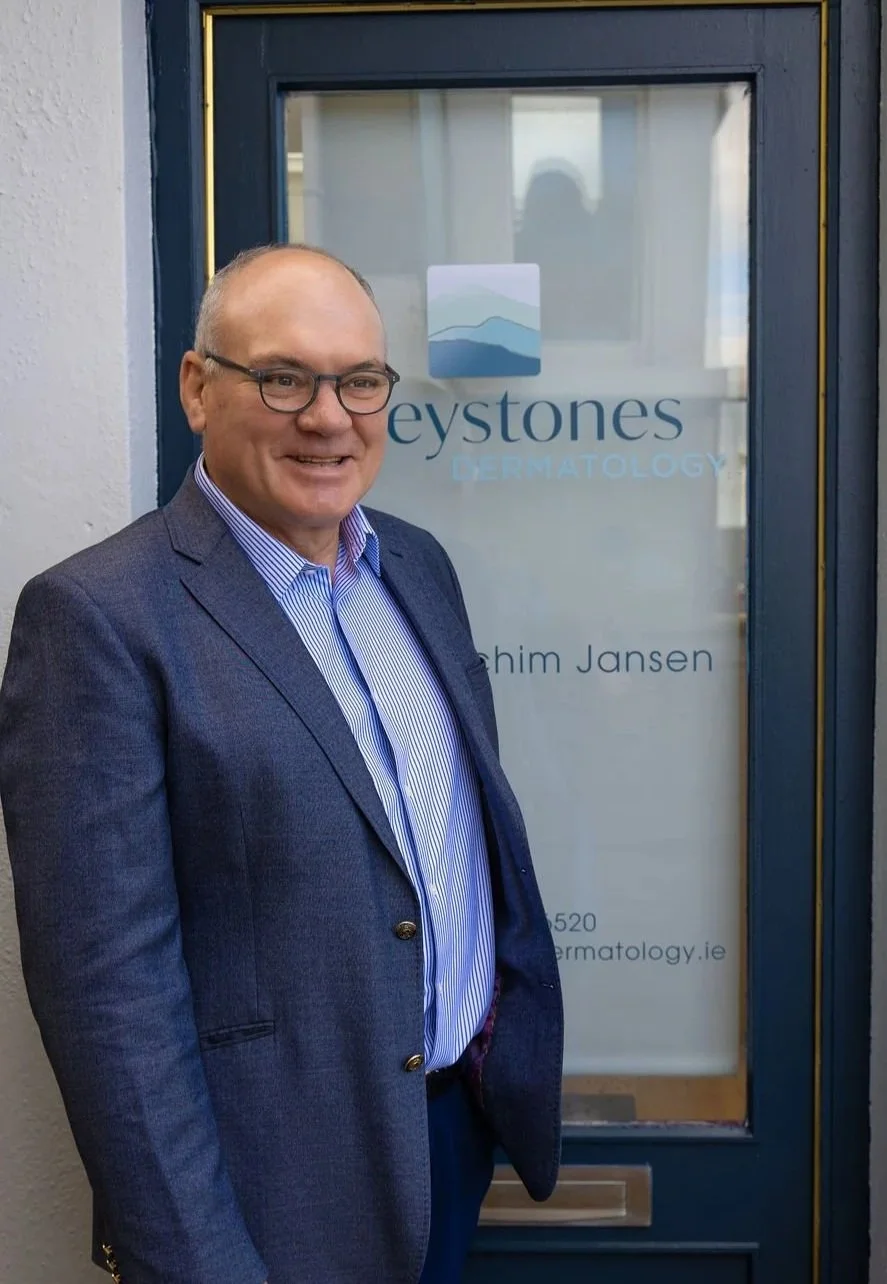 A smiling man in a blue blazer and glasses standing in front of a door with a sign that reads 'Keystones Dermatology' and 'Joachim Jansen'.