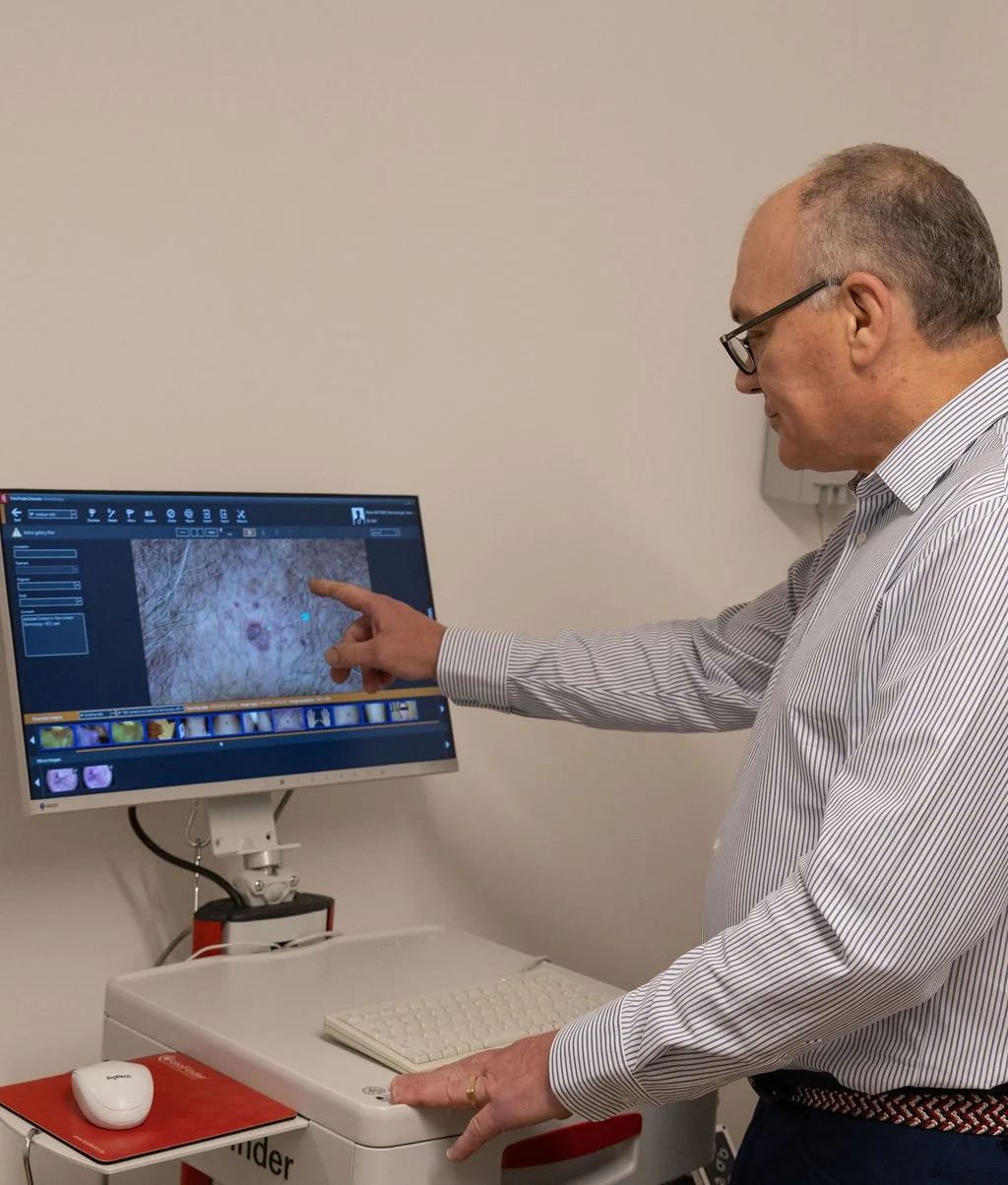 A man in a suit and glasses points at a medical image on a computer monitor, which displays a close-up view of skin or tissue.