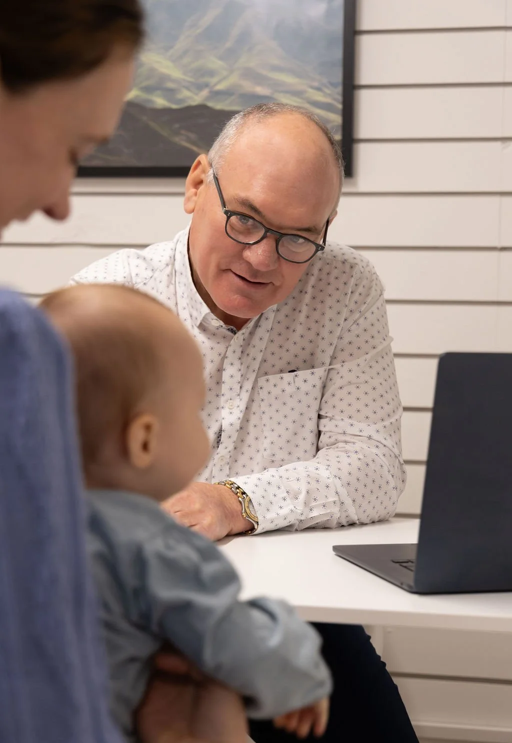 A man with glasses and a white patterned shirt talking to a woman holding a baby in a room with white horizontal panel walls and a landscape painting on the wall.