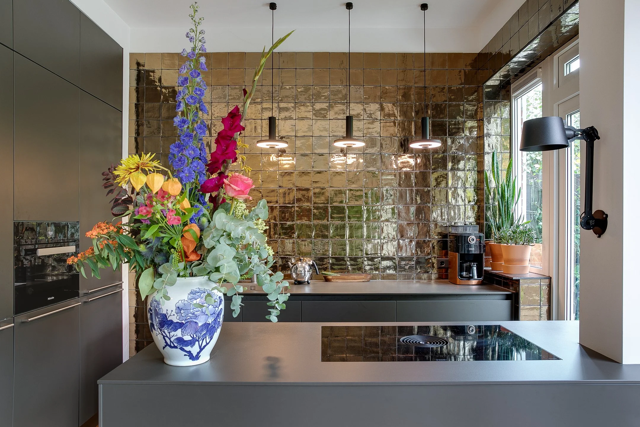Interieur van een moderne bulthaup keuken met goudkleurige tegelwand, zwarte hanglampen en een kleurrijk bloemboeket