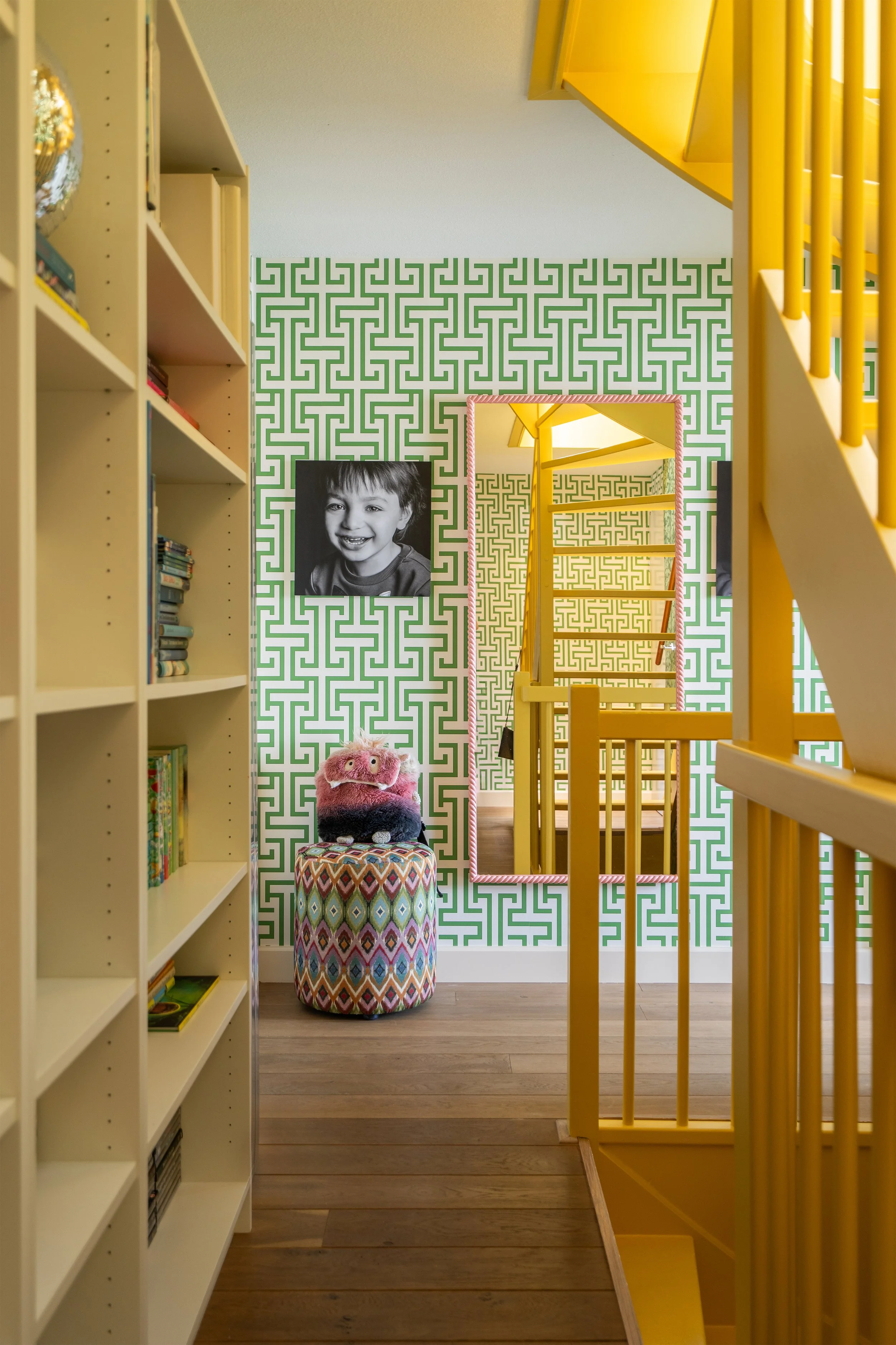 Kleurrijke overloop met een grafisch Thibaut Ming Trail behang, een Claudi poef en spiegel, met zicht op een gele Little Greene Giallo trap.