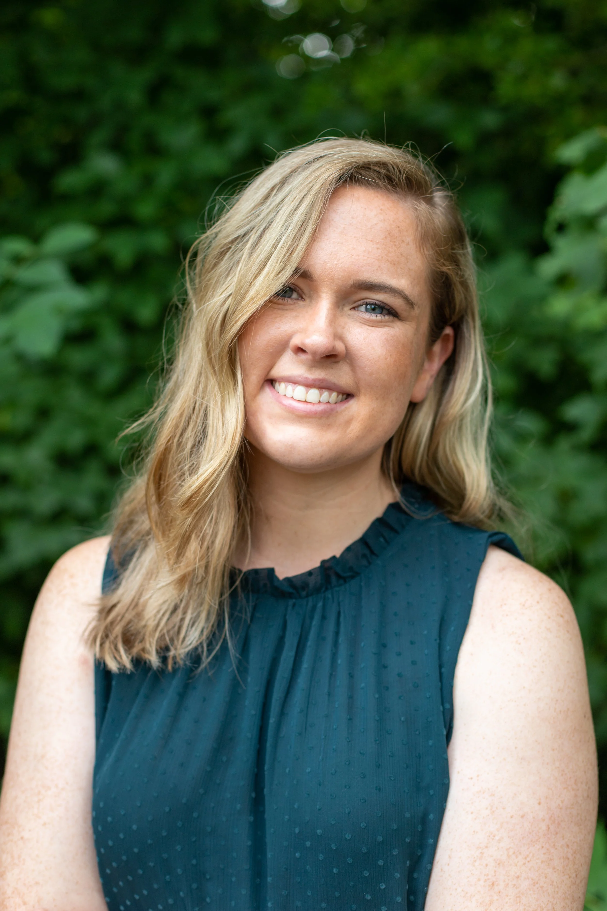 A smiling woman with long blonde hair, blue eyes, and freckles, wearing a sleeveless dark blue dress with ruffle trim and small polka dot pattern, outdoors in front of green foliage.