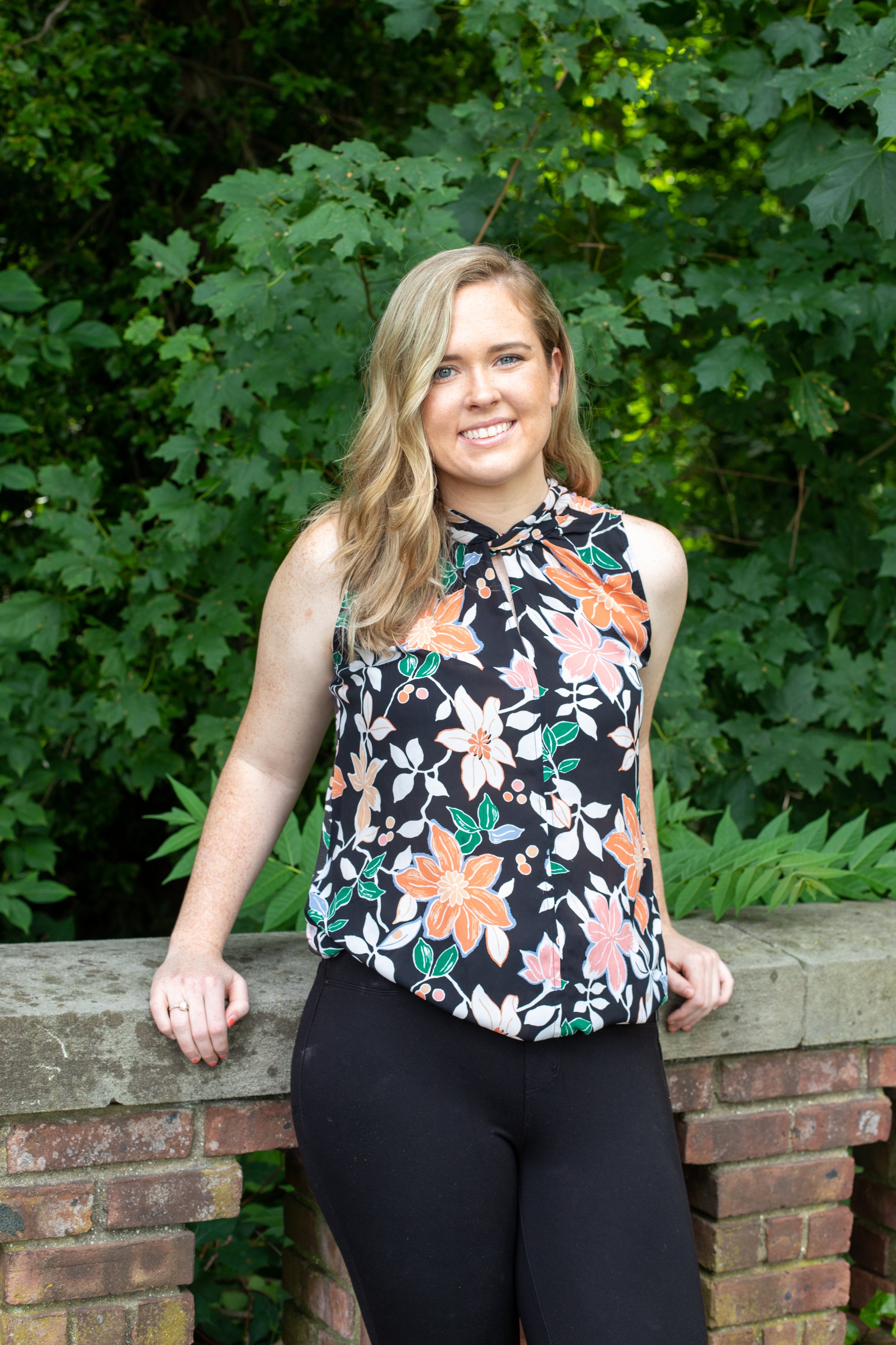A young woman with blonde hair and blue eyes standing outdoors in front of green leafy bushes. She is wearing a sleeveless, floral-patterned blouse and black pants, leaning against a low brick wall.