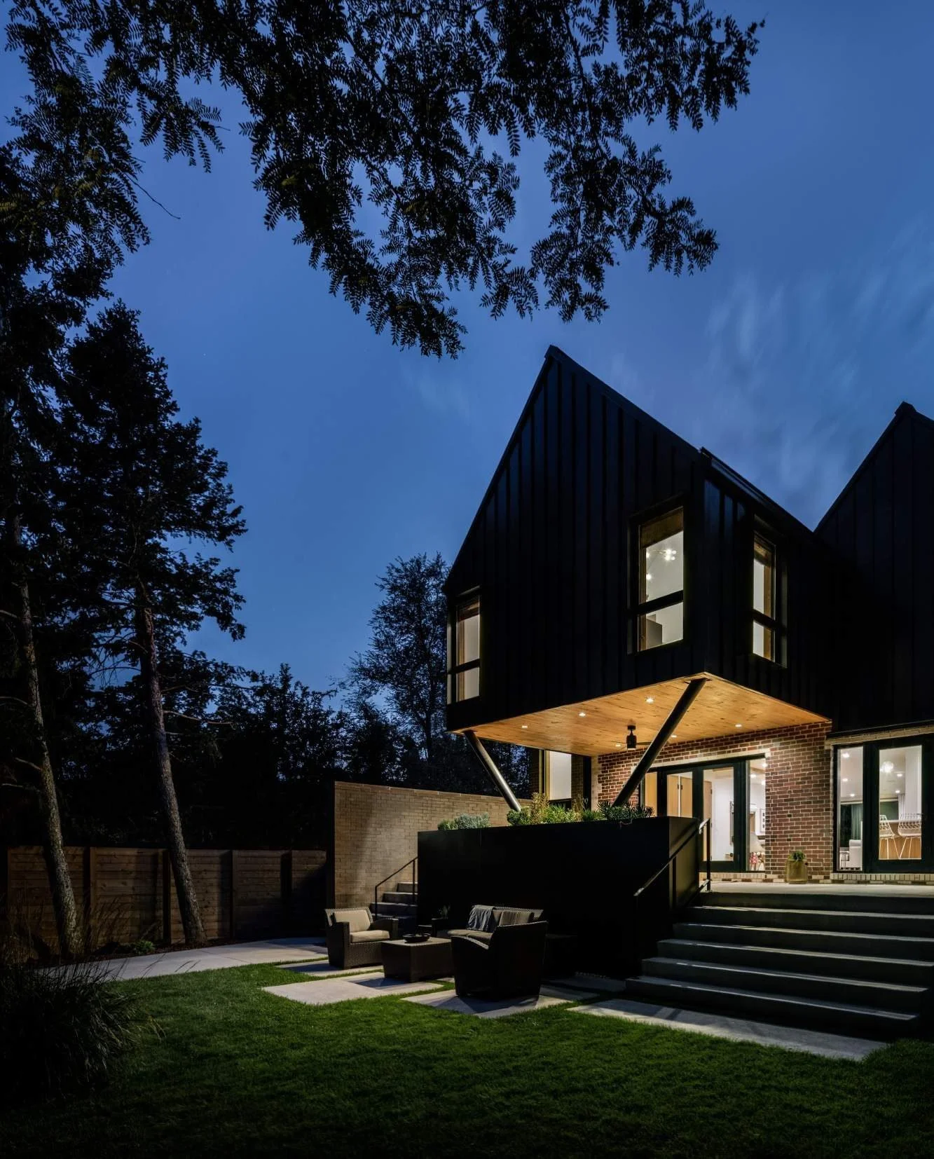 The cantilevered gable, lit from beneath, shelters the rear yard at dusk.