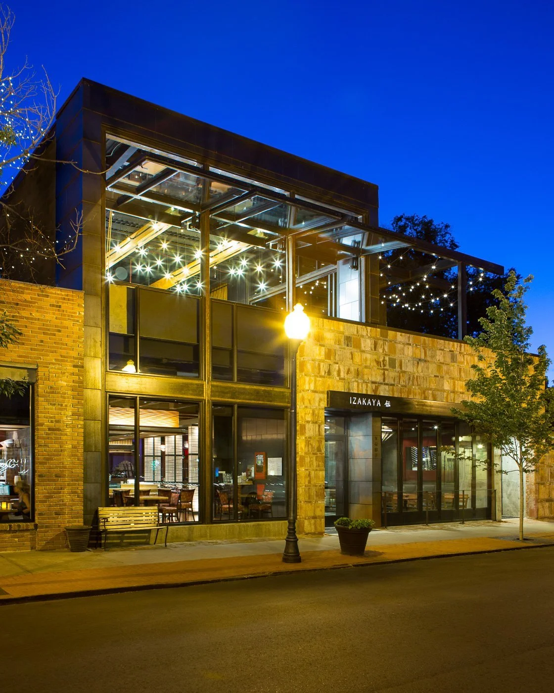 The corner at dusk — weathered steel, clay tile, and stone against the lit interior.
