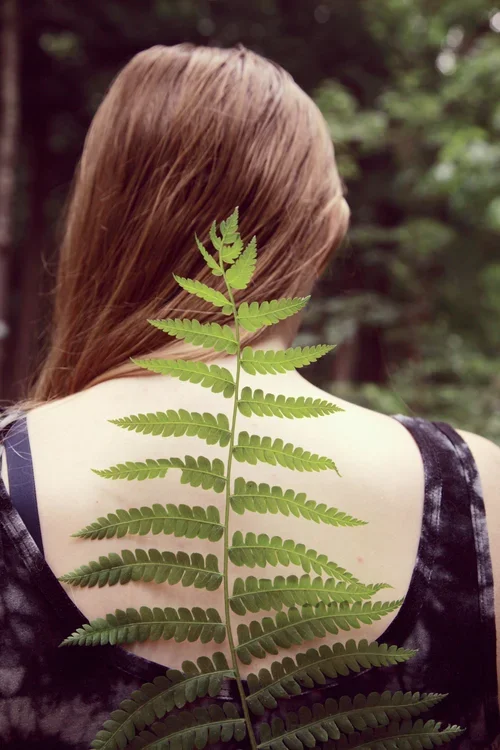 A woman with long hair facing away outdoors, with a large fern leaf covering her back.