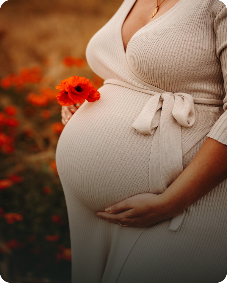 Pregnant woman in beige ribbed dress holding her belly with one hand and a red flower with the other, standing outdoors.