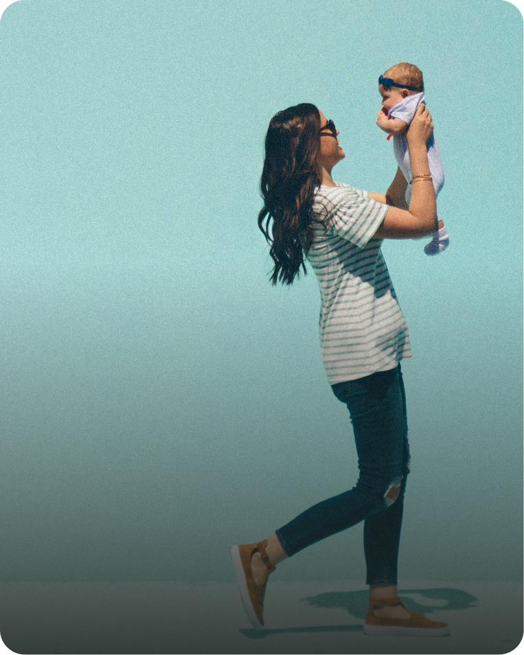 A woman with long dark hair, wearing sunglasses, a striped t-shirt, ripped jeans, and tan sneakers, holding a smiling baby with a blue headband up in the air against a plain blue background.
