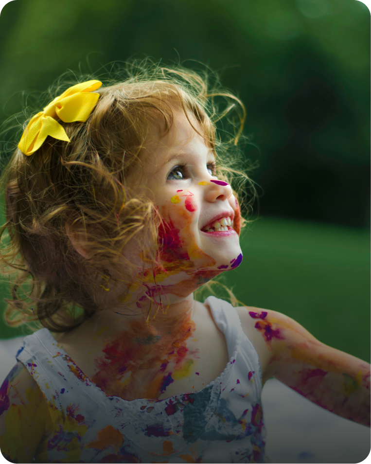 A young girl with curly red hair, wearing a yellow bow, smiling outdoors with paint on her face and arms.