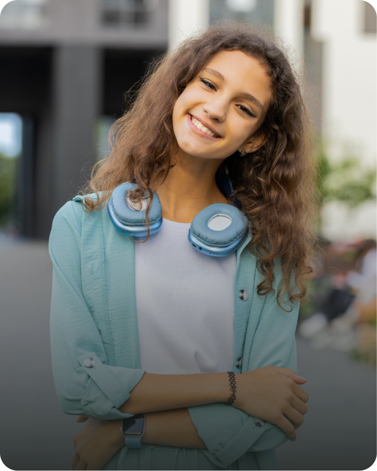 Young woman smiling outdoors with headphones around her neck, wearing a light blue jacket and white shirt.