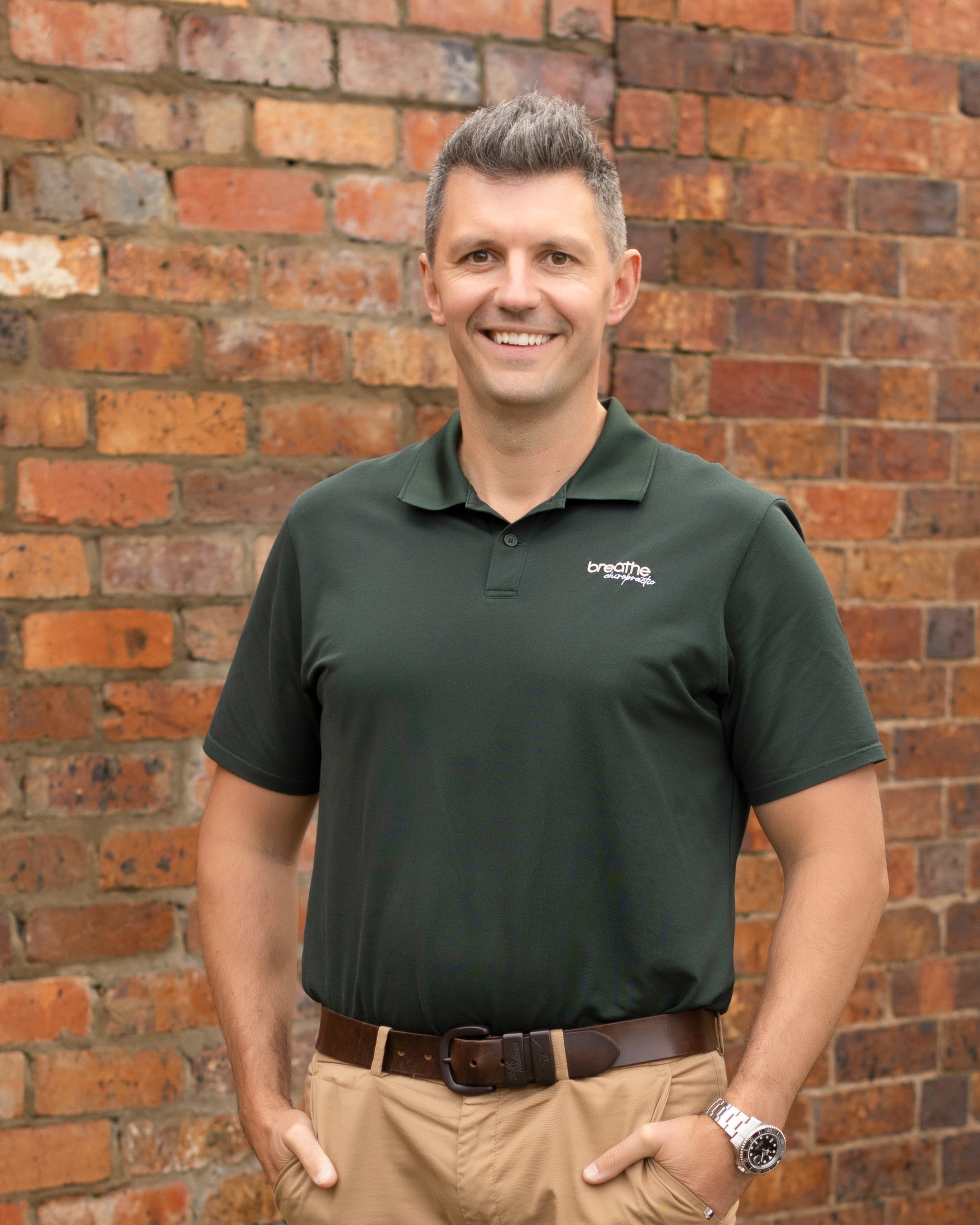 A smiling man wearing a dark green polo shirt with a small logo, beige pants, a brown belt, and a silver watch standing against a red brick wall.