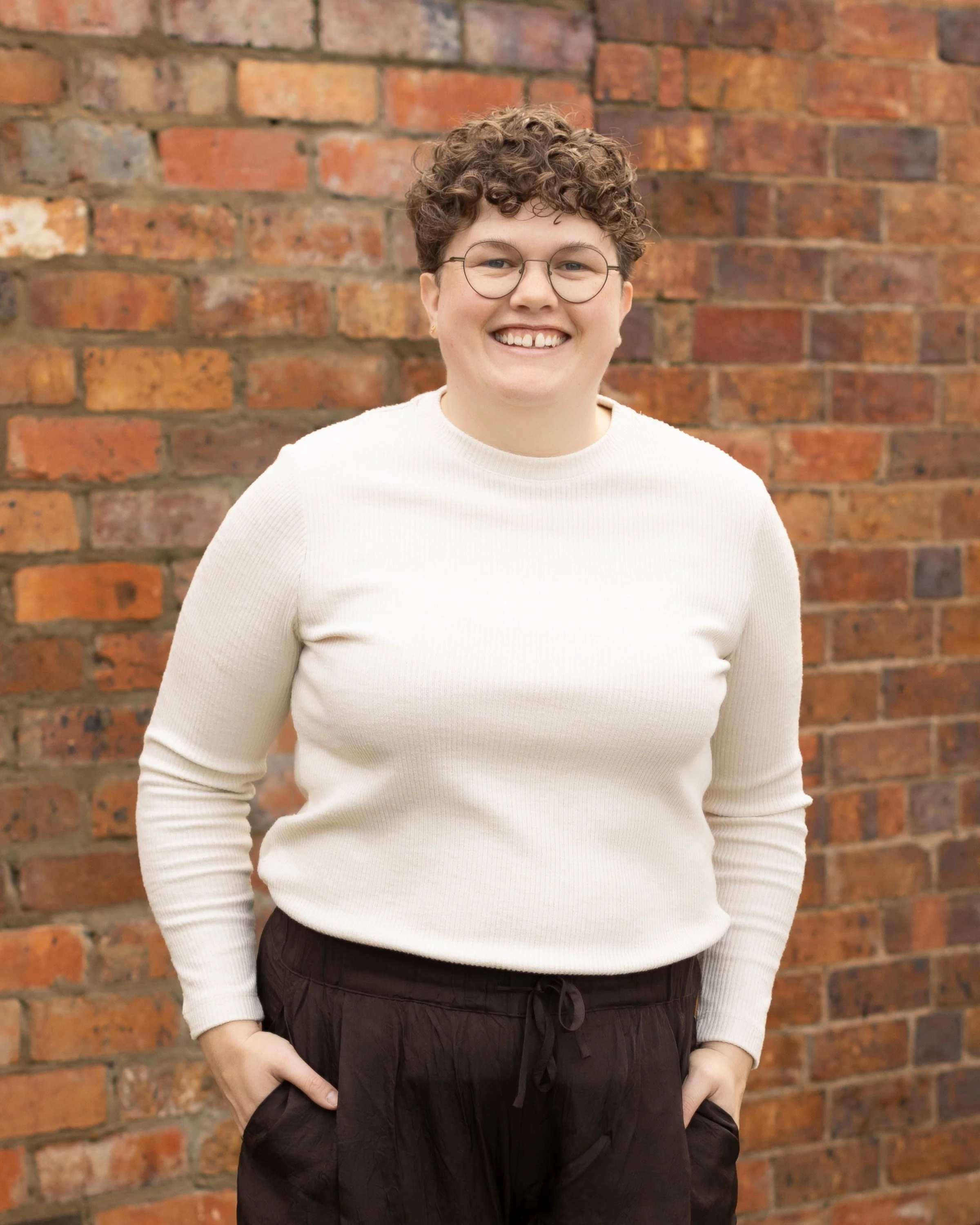 A person with short, curly hair wearing glasses, a white long-sleeve shirt, and dark pants, standing in front of a brick wall, smiling at the camera.