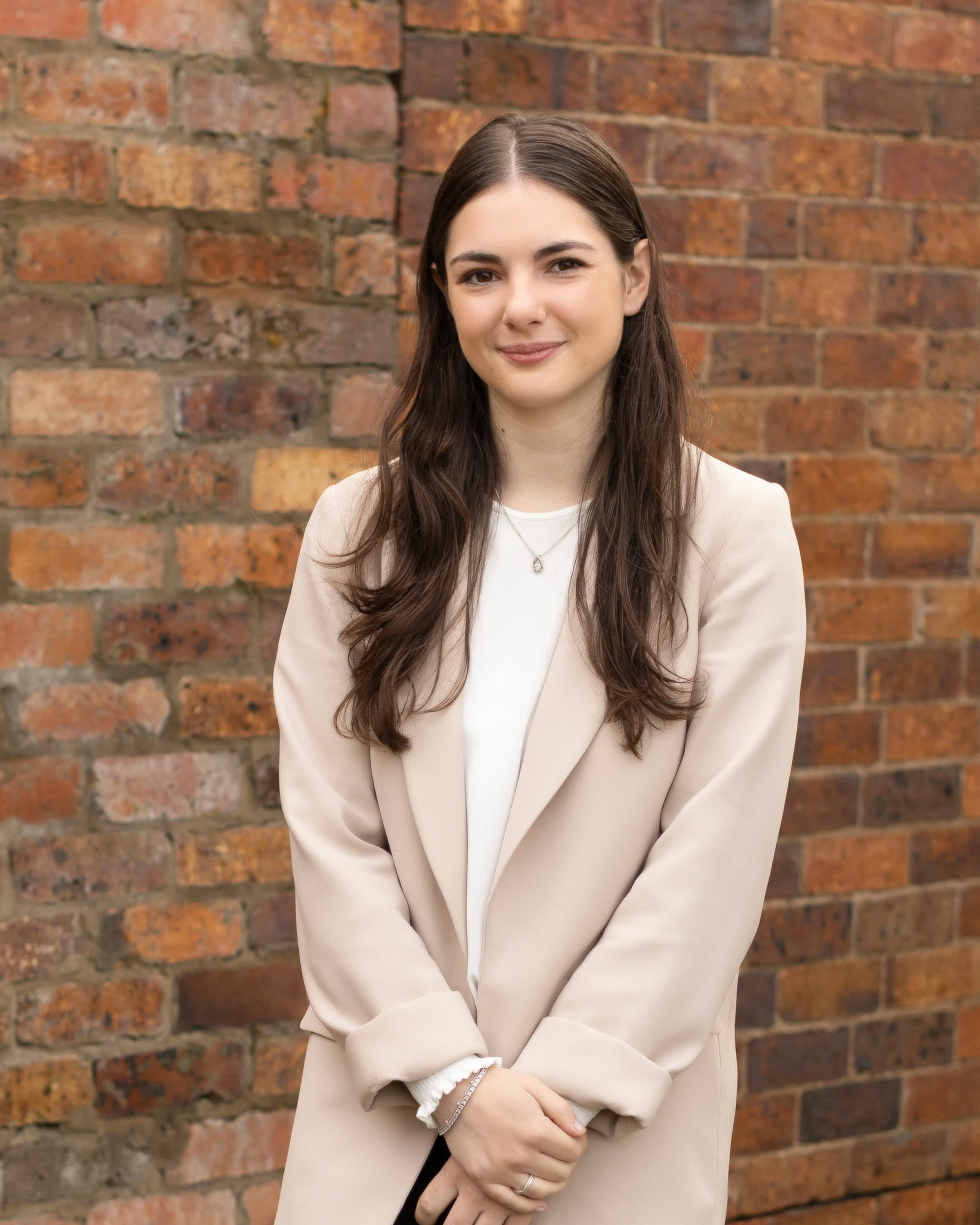 A young woman with long, dark hair and a gentle smile standing in front of a brick wall, wearing a light beige blazer over a white top and a silver necklace.