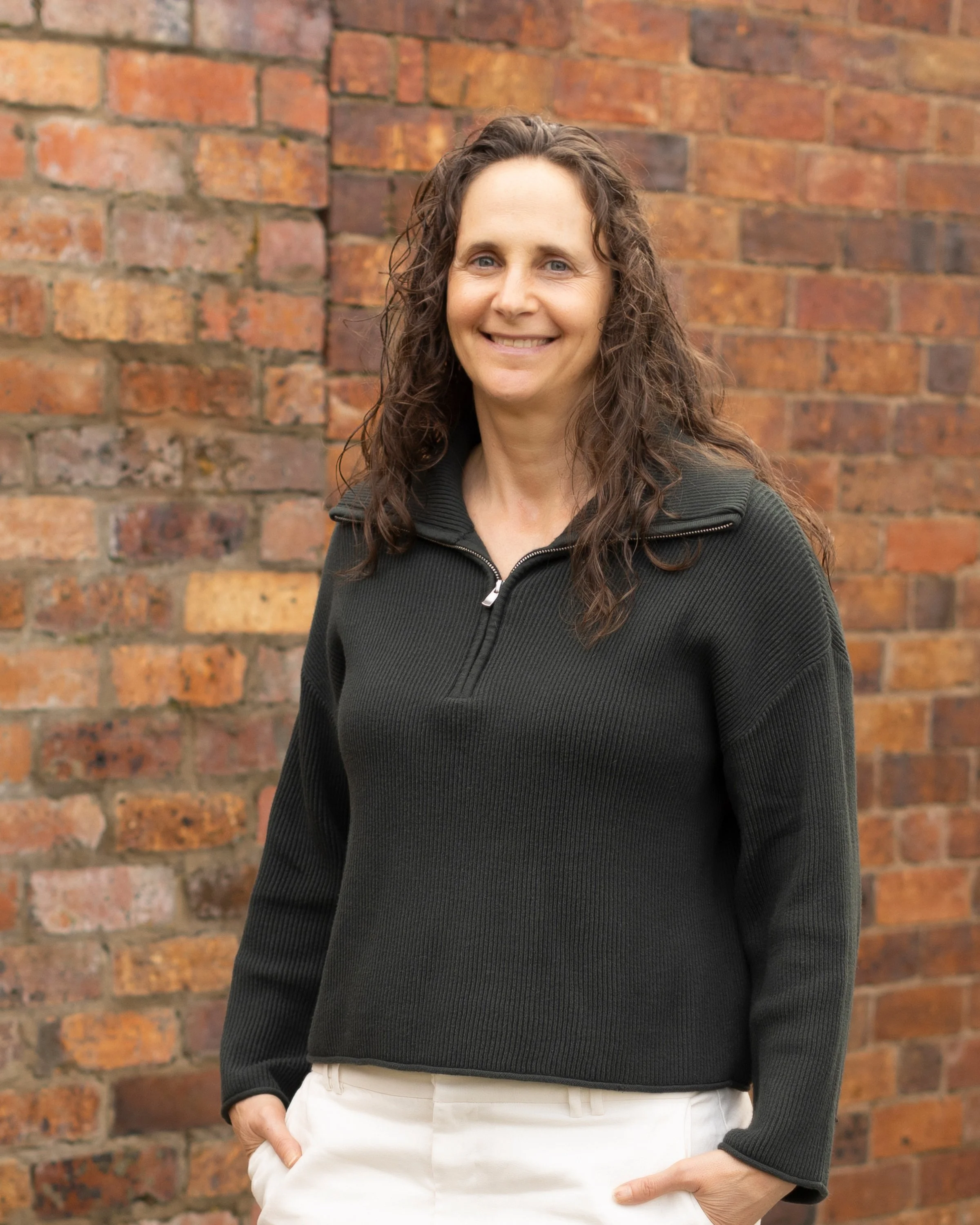 A woman with curly brown hair smiling, wearing a black zip-up jacket and white pants, standing in front of a brick wall.