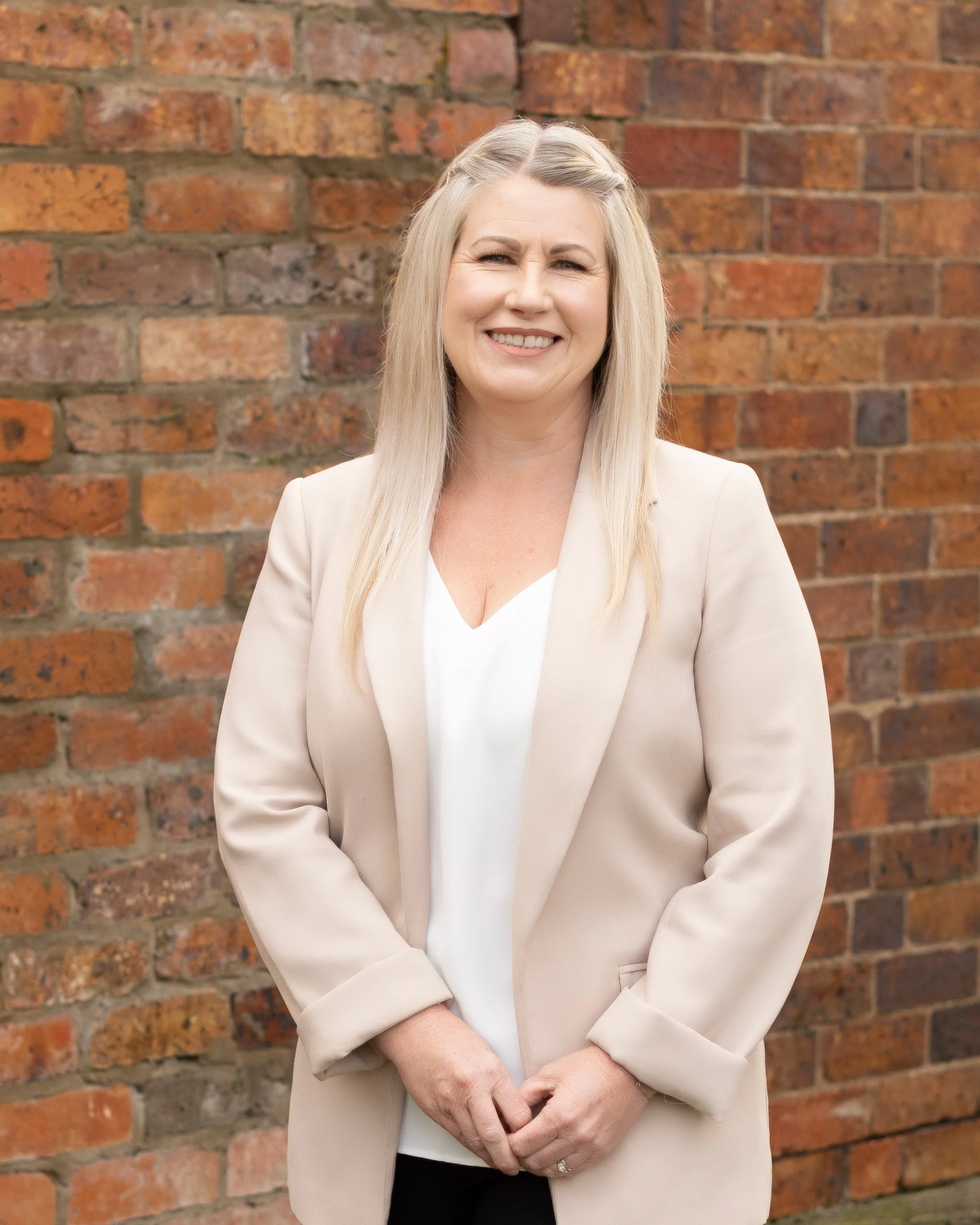 A woman with long blonde hair wearing a beige blazer and white top, standing in front of a red brick wall, smiling and holding her hands together in front of her.