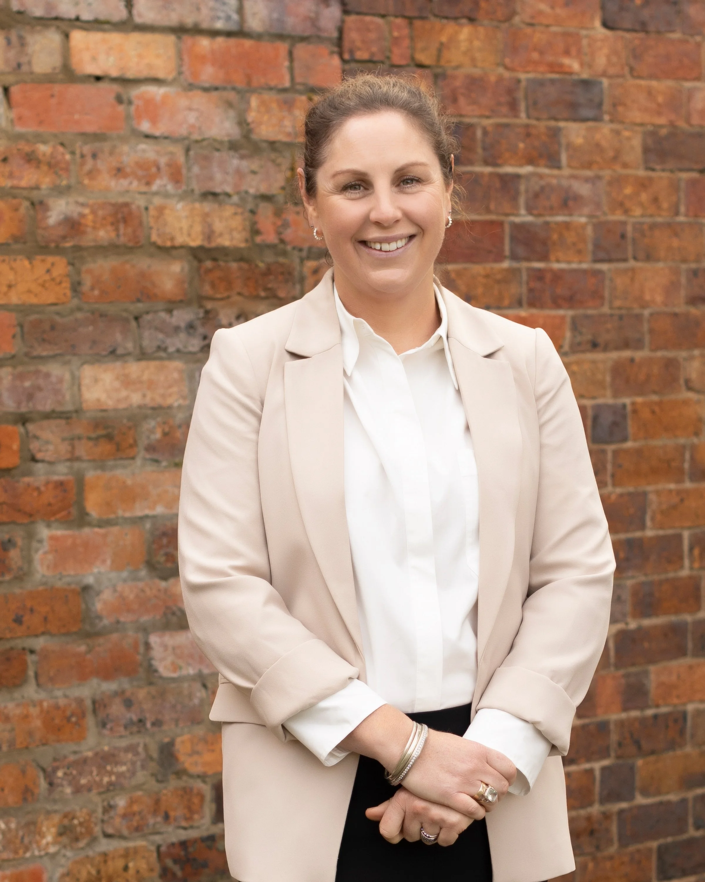 A smiling woman with light skin and brown hair tied back, wearing a beige blazer over a white shirt, standing in front of a brick wall.
