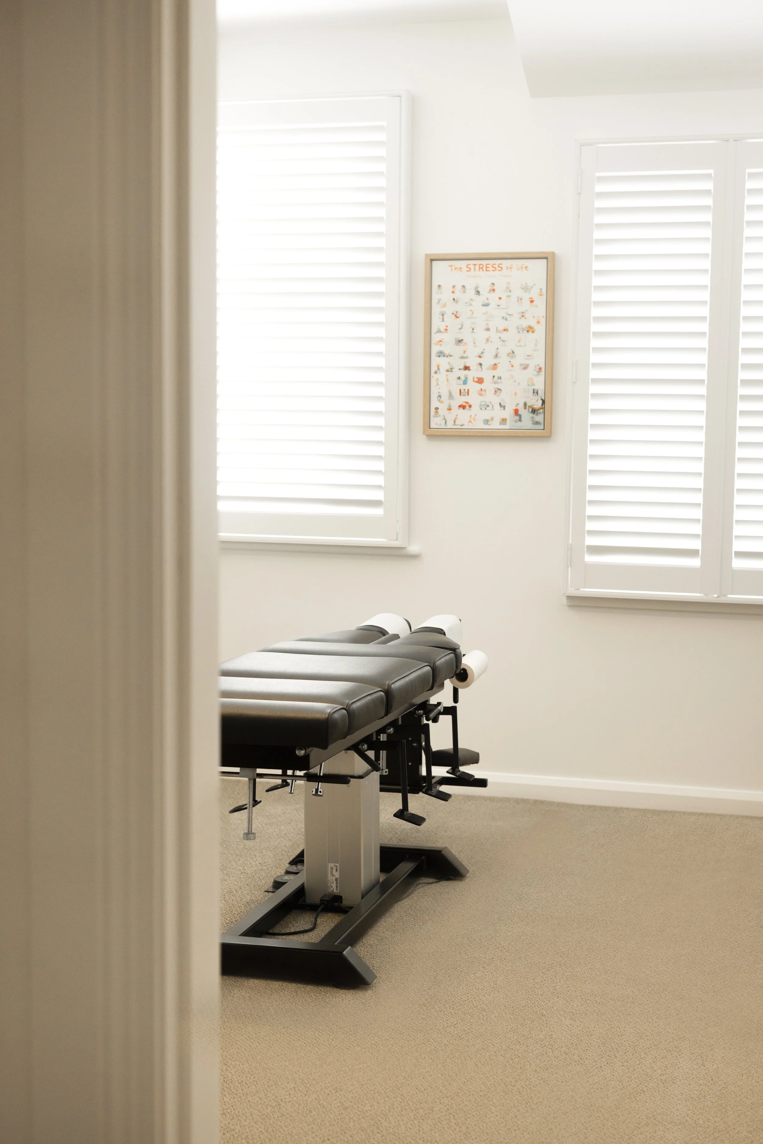 An empty examination or treatment room with a medical or chiropractic chair, a framed poster on the wall reading 'The Stress of life,' and white blinds covering windows. The room has beige carpet and white walls.