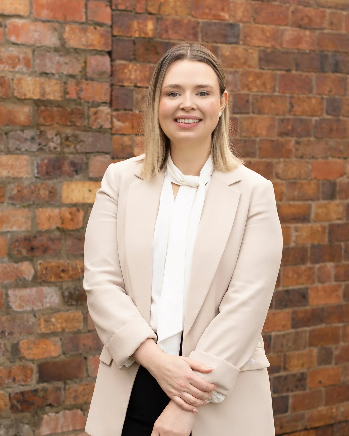 A woman wearing a white blouse and beige blazer standing in front of a brick wall, smiling with her arms crossed.