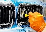 Close-up of a person cleaning a car's front grille with a yellow sponge, wearing an orange glove.