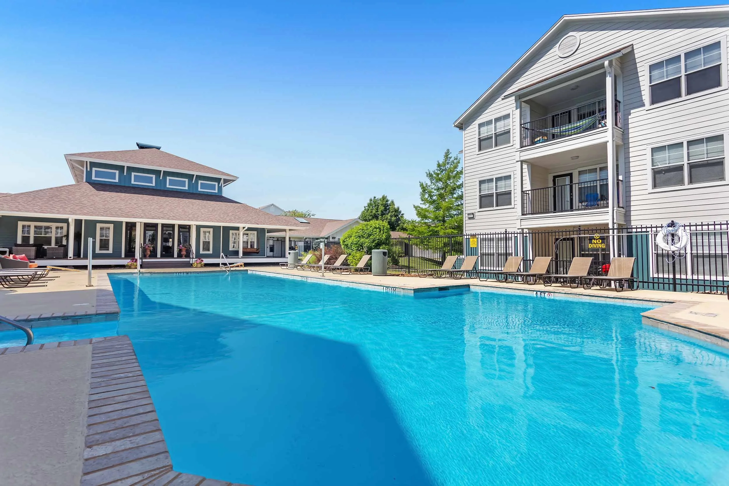 Image of a swimming pool area at an apartment complex with blue sky, poolside lounge chairs, a clubhouse, and a multi-story residential building with balconies.