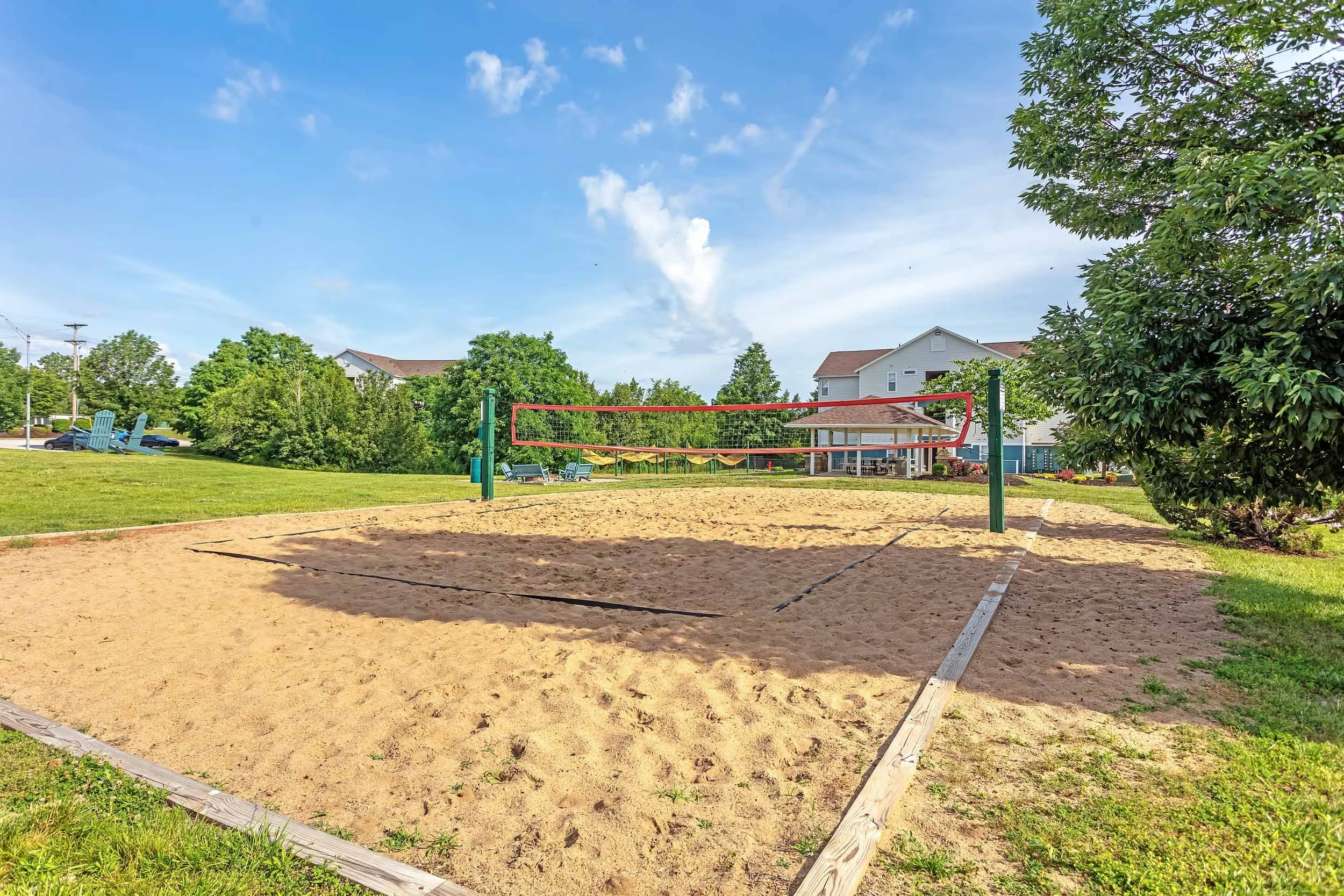 A sand volleyball court with a net set up in a park. Green grass surrounds the court, and there are trees, houses, and a clear blue sky in the background.