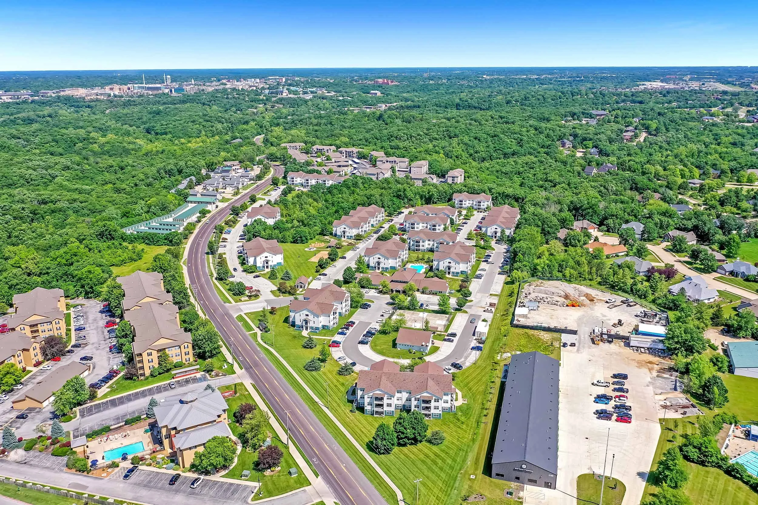 Aerial view of a suburban area with green trees, residential buildings, parking lots, a swimming pool, and a road cutting through the neighborhood, with a city skyline and more greenery in the background.