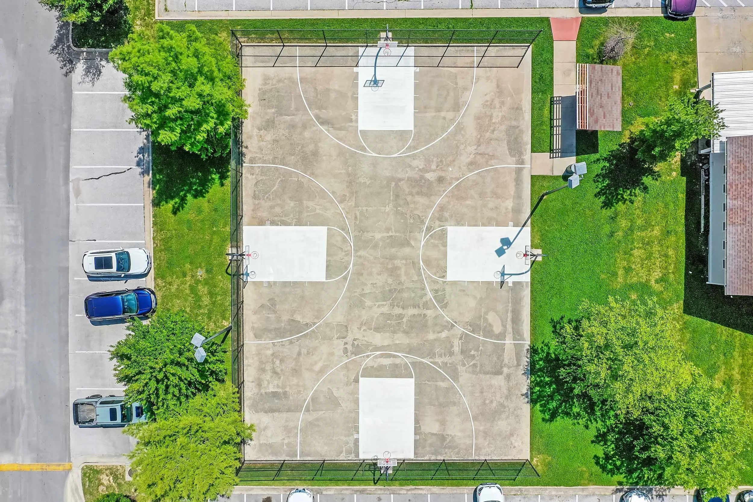 An aerial view of an outdoor basketball court with four basketball hoops, surrounded by parked cars, trees, and grassy areas.