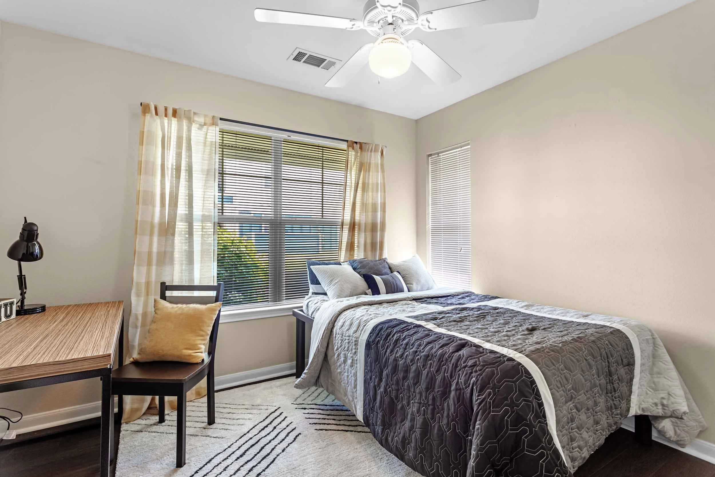 A bedroom with a bed, a window with blinds and curtains, a ceiling fan with lights, a desk with a chair, a lamp, and decorative pillows.