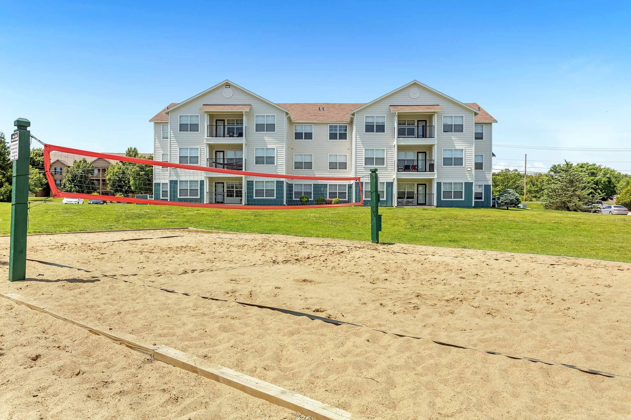 A sandy beach volleyball court with a red net, set against a background of a modern four-story apartment building with balconies and large windows, surrounded by greenery and a parking lot.