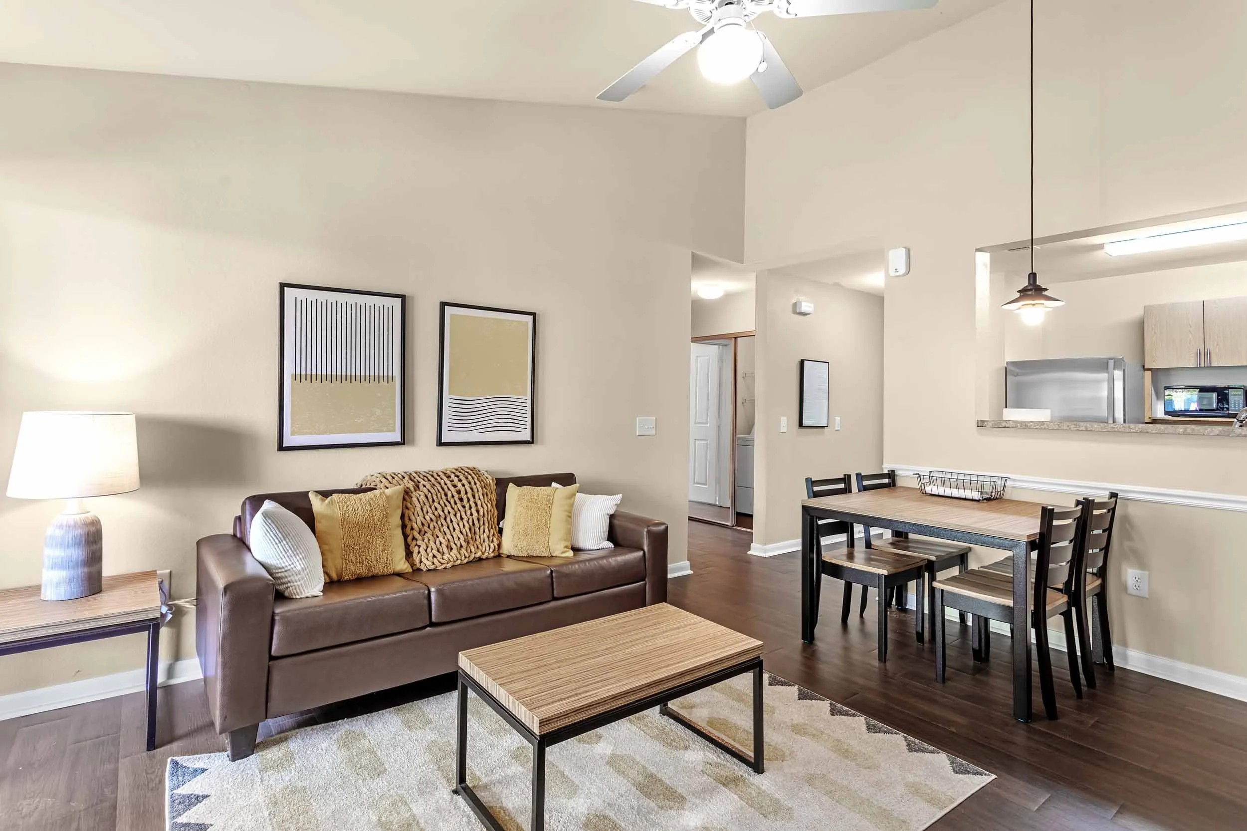 Living room with beige walls, a brown leather sofa with yellow and white pillows, a wooden coffee table, a side table with a lamp, two framed abstract art pieces on the wall, and a dining area with a rectangular table and six chairs. The kitchen is visible in the background with a stainless steel refrigerator, microwave, cabinetry, and a countertop. There are ceiling fans and hanging light fixtures.