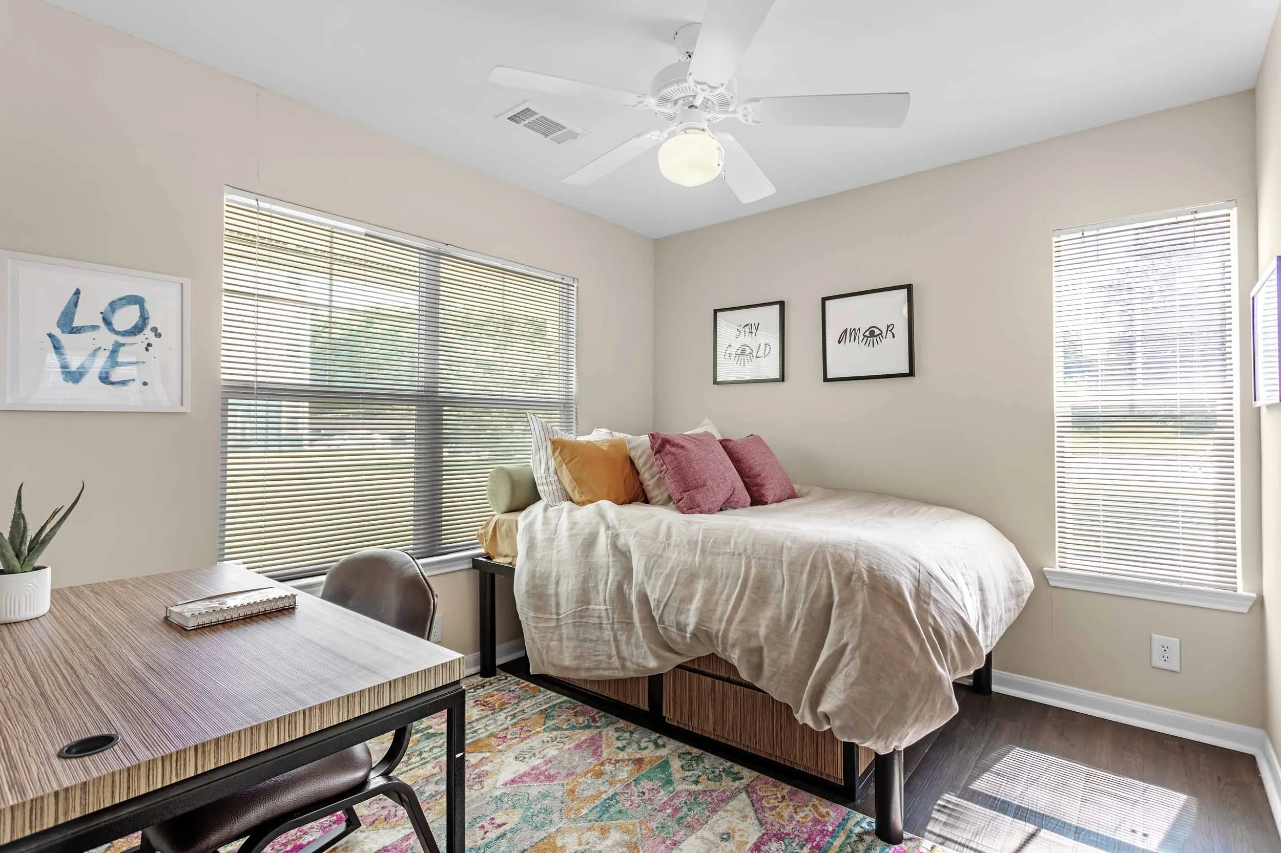 A bedroom with a bed against a wall with two framed prints, one says "Stay Gold" and the other says "Amor" with eye illustrations. The room has large windows with blinds, a ceiling fan with a light, a desk with a notebook, and a colorful area rug.