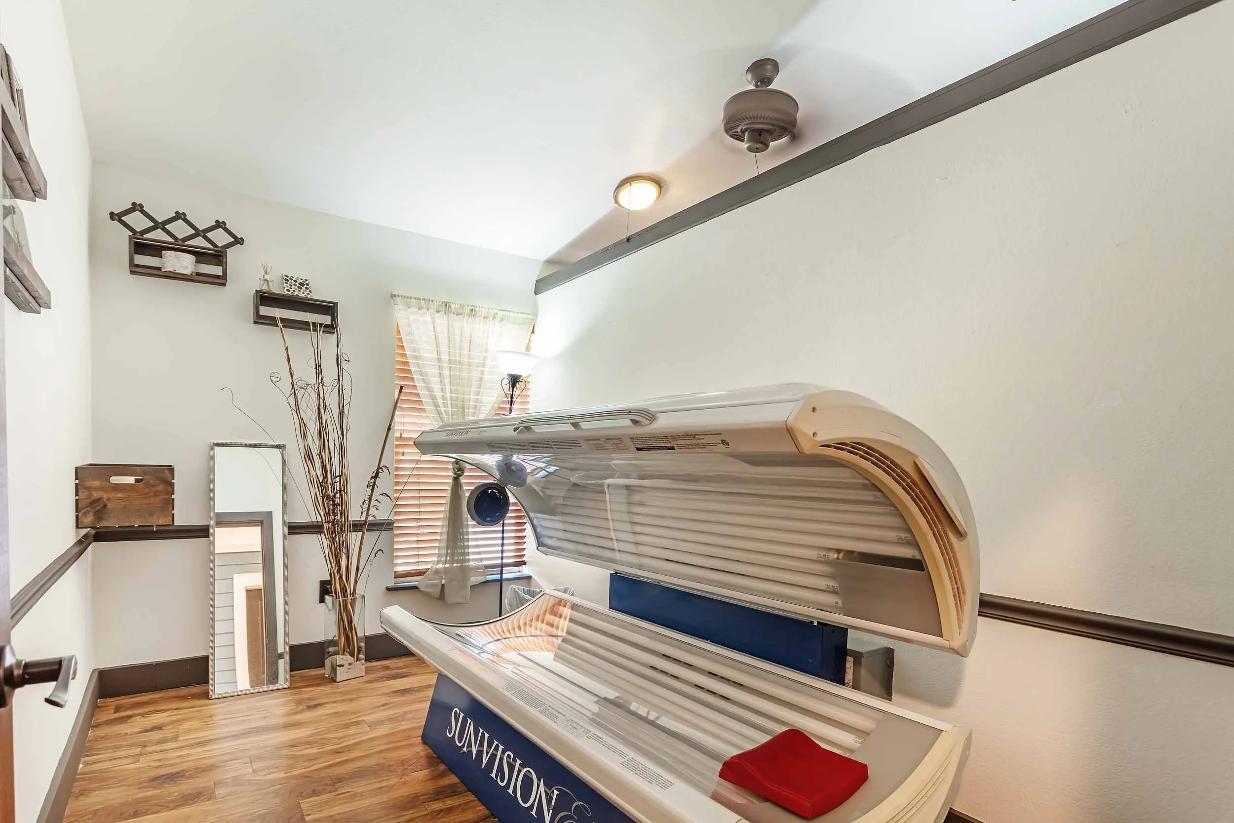 Tanning room featuring a decompression table with a beige cushioned surface and a blue base, a window with wooden blinds, a tall floor lamp, a mirror leaning against the wall, wall-mounted shelves, decorative branches, and a ceiling fan with a light.