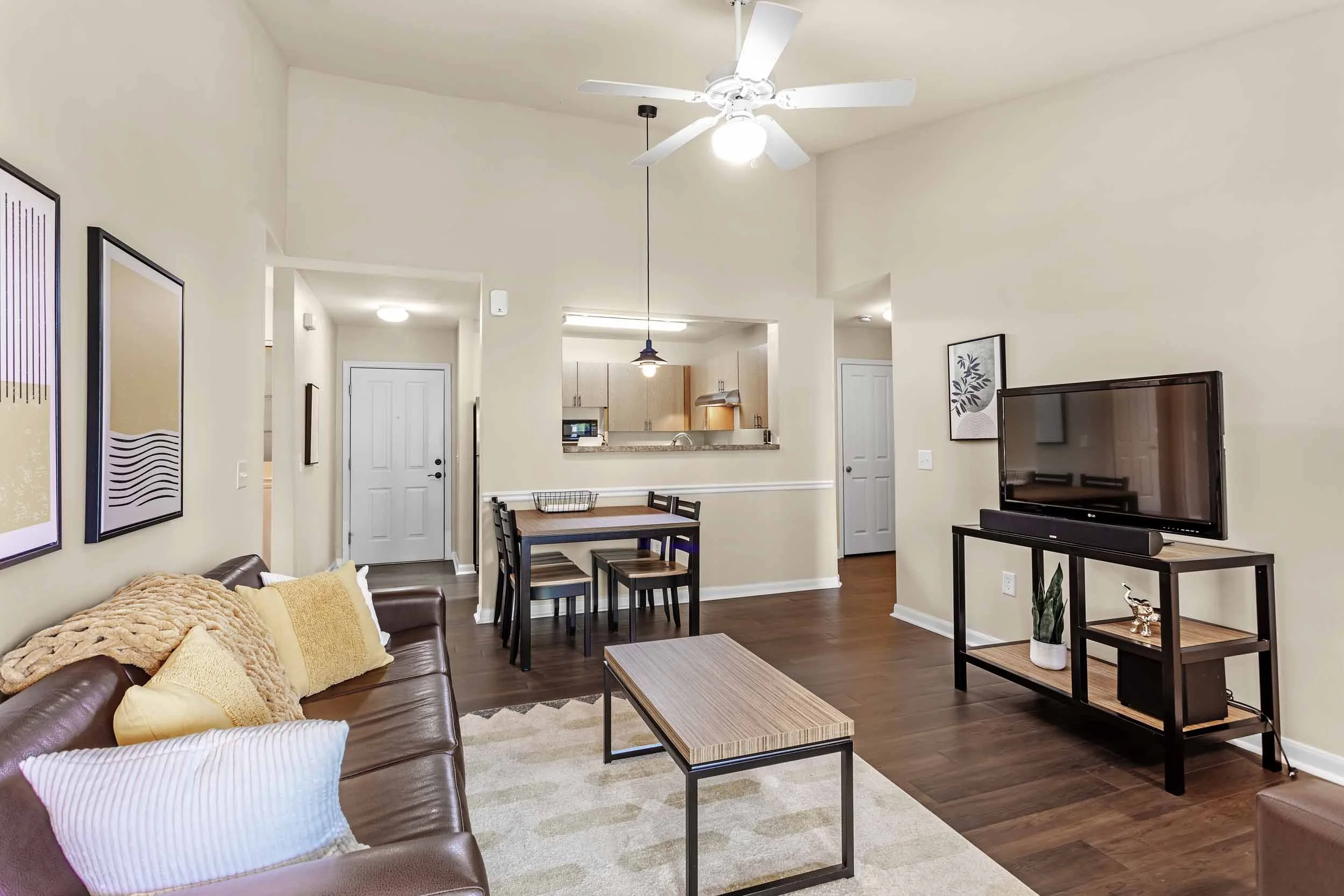 Living room with a brown leather sofa adorned with yellow and white pillows, a patterned area rug, a wooden coffee table, and a TV on a modern black stand, with artwork on the walls and a ceiling fan.