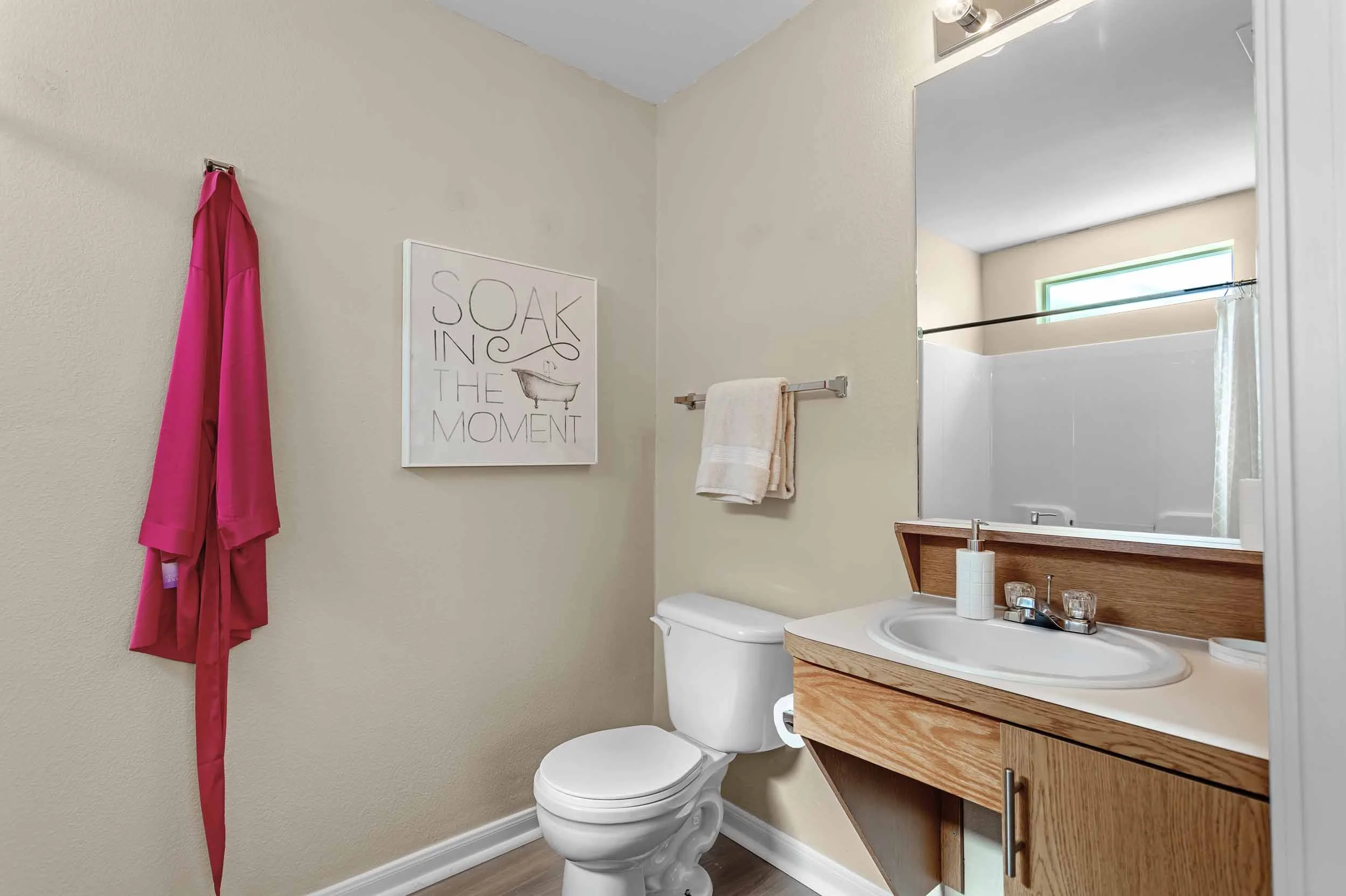 A small bathroom with a white toilet, wooden cabinet with a sink, mirror, towel rack, and a pink bathrobe hanging on the wall. There is a framed sign that says "Sow in the moment" and a small window with a shower curtain.