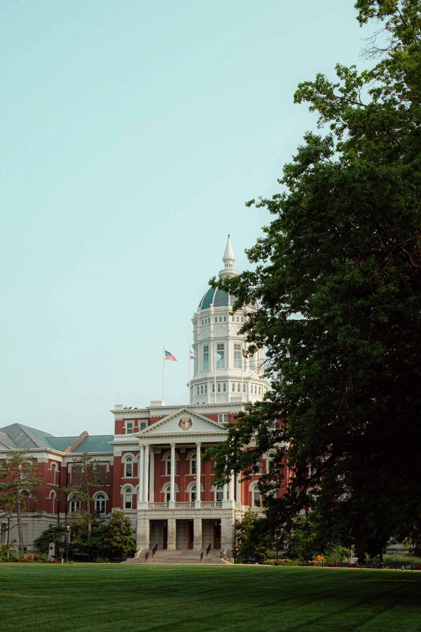 Mizzou building with a red-brick facade, white columns, and a dome topped with a spire, partially obscured by green trees and a clear sky.