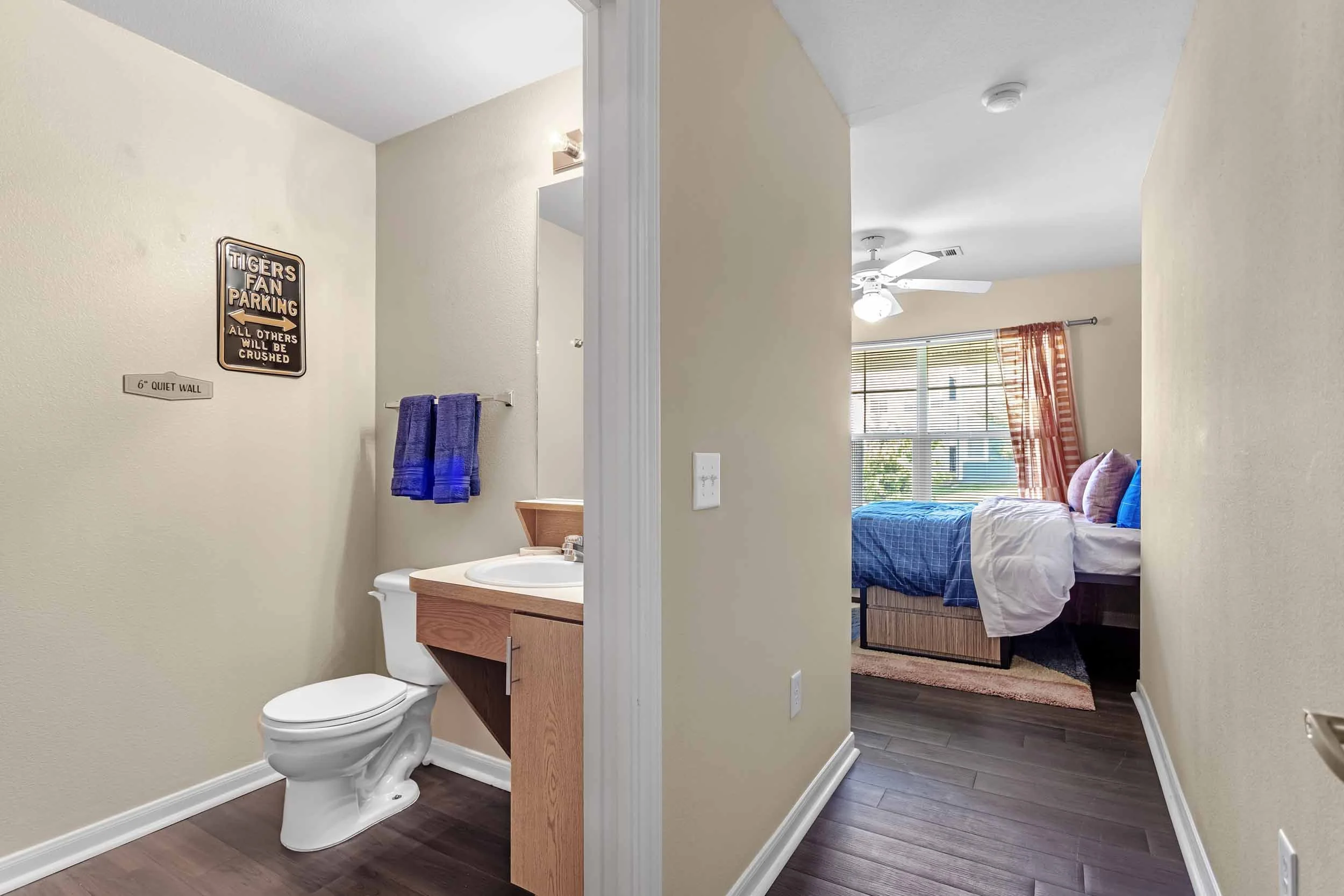 View of a bedroom with a large window, bed with blue bedding and purple pillows, and a ceiling fan. Adjacent bathroom with a toilet, a wooden vanity with a sink, purple towels, and decorative signs on the wall.