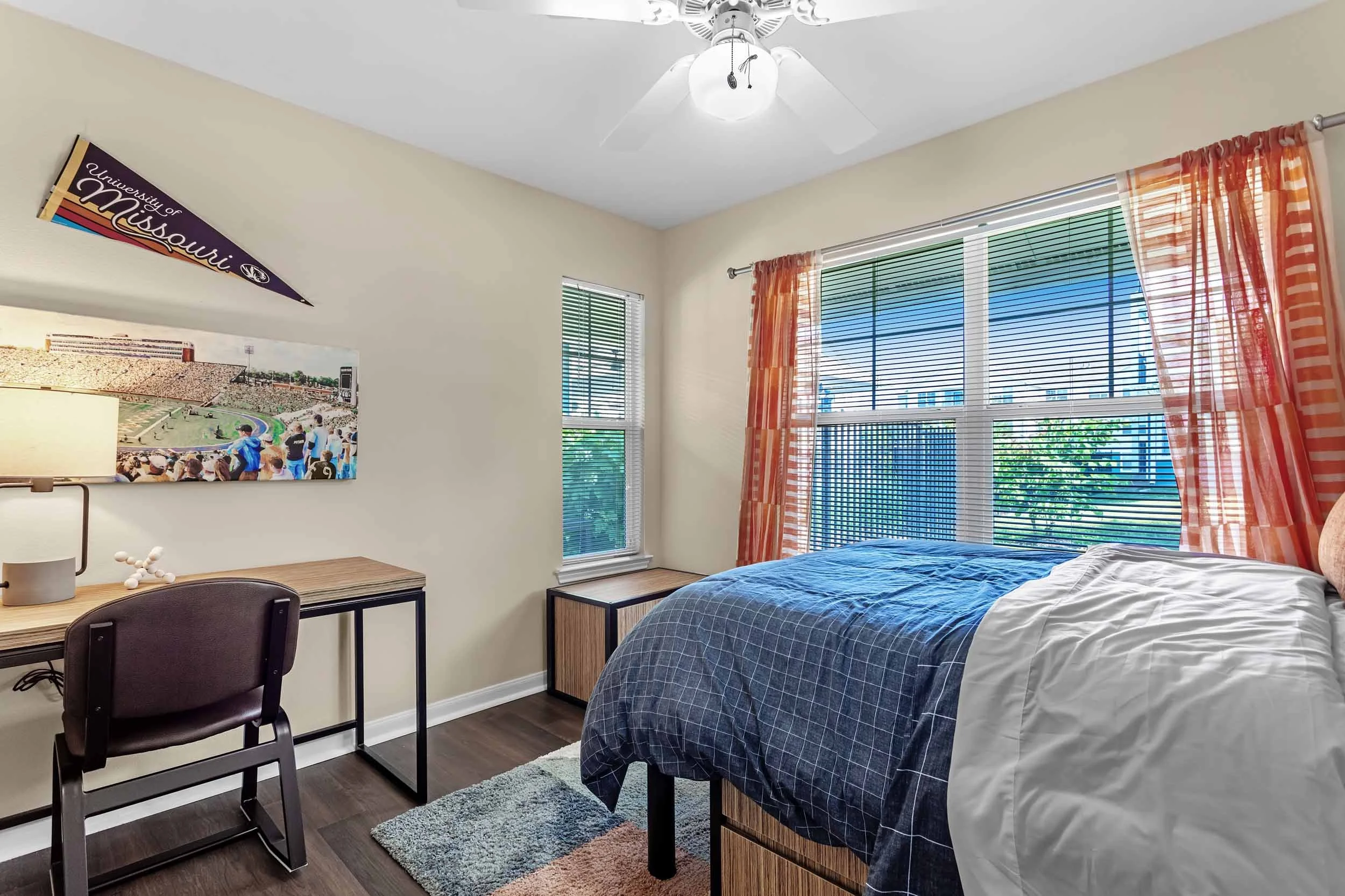 A bedroom with a bed, large windows with blinds and orange curtains, a ceiling fan, a desk with a chair, a wall art poster, and sunlight coming through the windows.