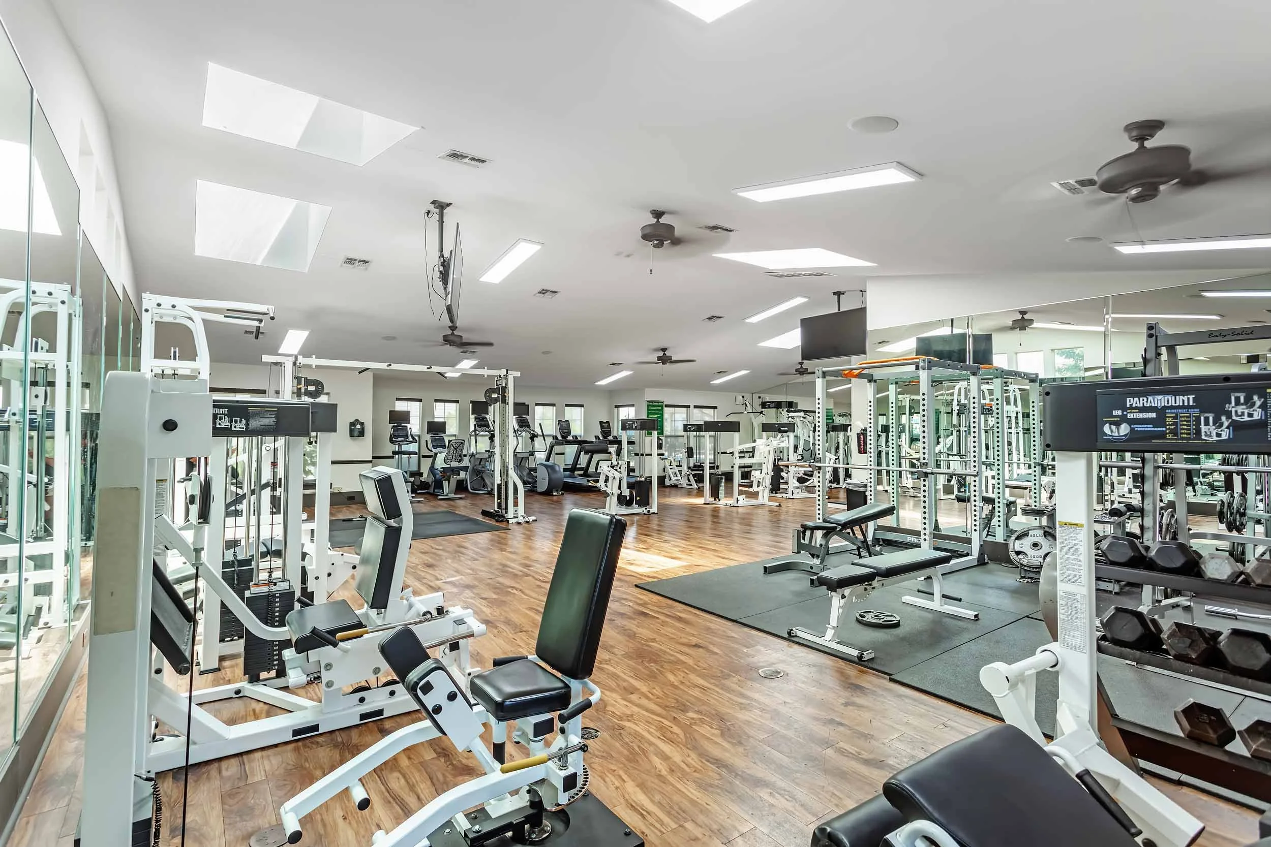Inside a well-lit fitness gym with various workout machines, free weights, and exercise benches, hardwood floors, ceiling fans, and skylights.
