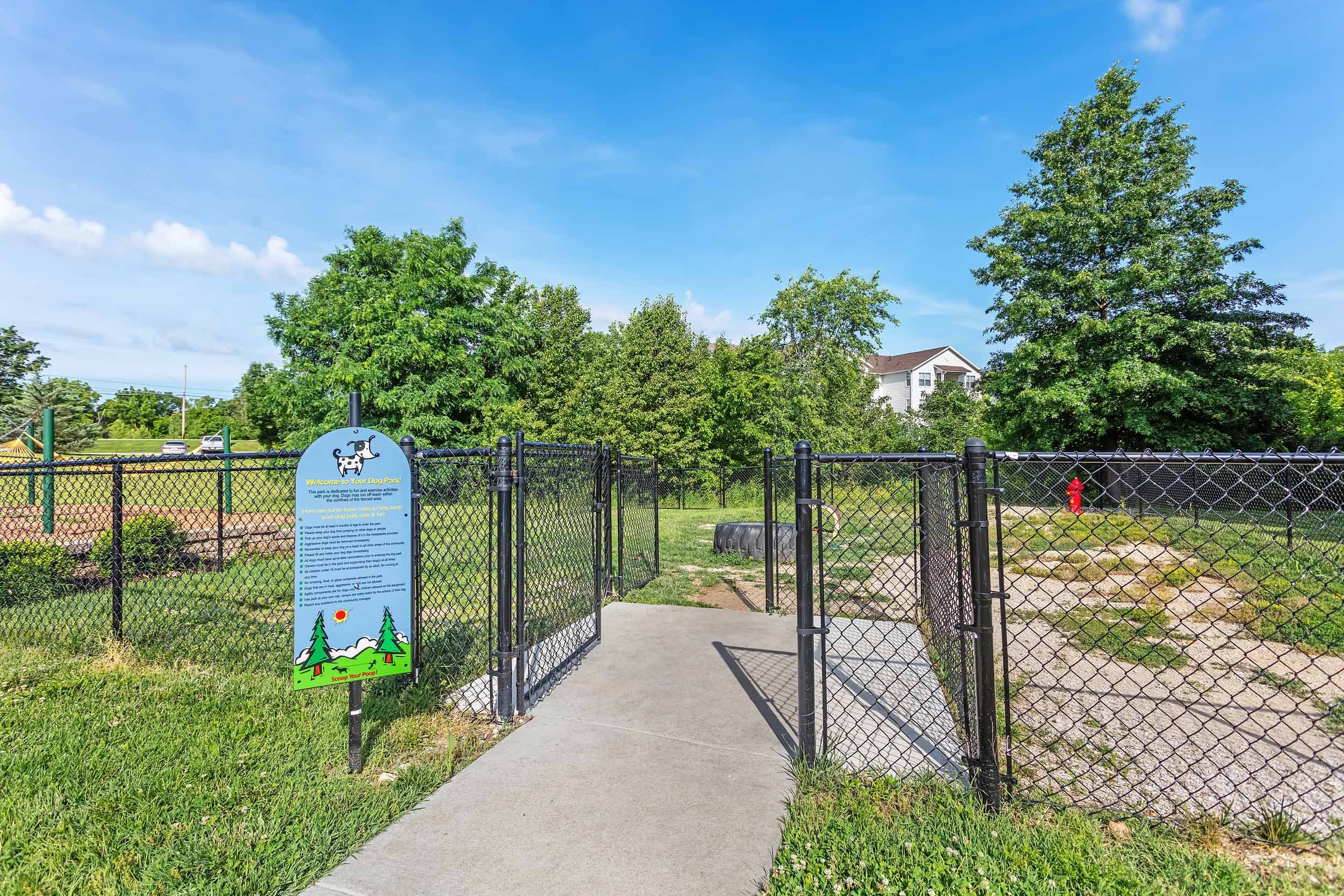 Entrance to a dog park with a black metal gate and a sign that says 'Welcome to Your Dog Park!'. The park is surrounded by a black metal fence, with lush green grass and trees, and a sidewalk leading inside. There are some residential buildings visib