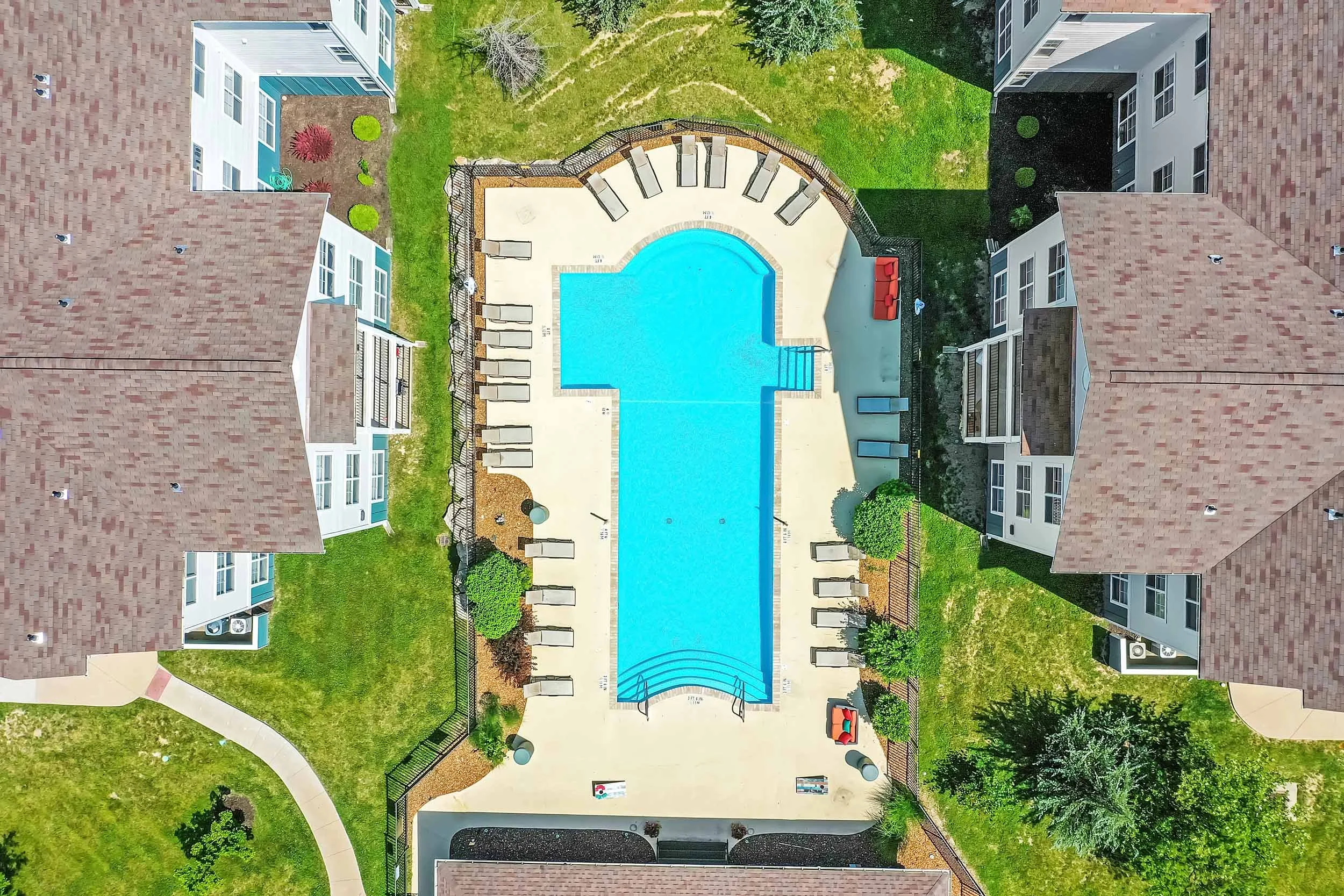An aerial view of an apartment complex outdoor pool area with lounge chairs, surrounded by green lawns, trees, pathways, and residential buildings with tiled roofs.