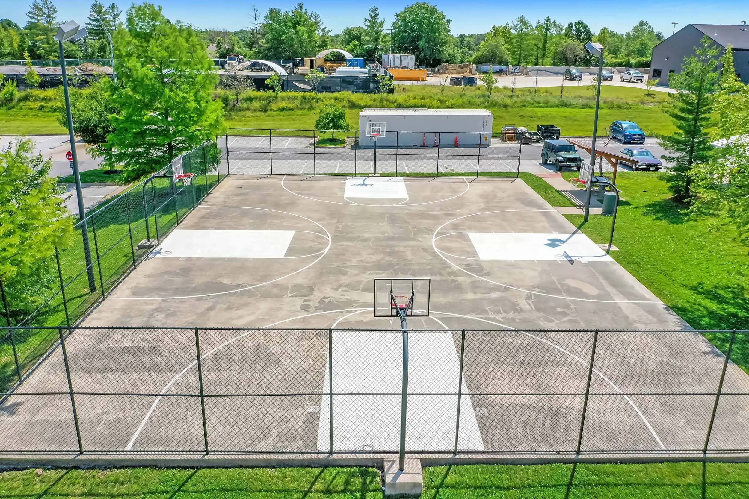 Empty outdoor basketball court with four hoops, surrounded by a safety fence, with parking lot and trees in the background on a sunny day.