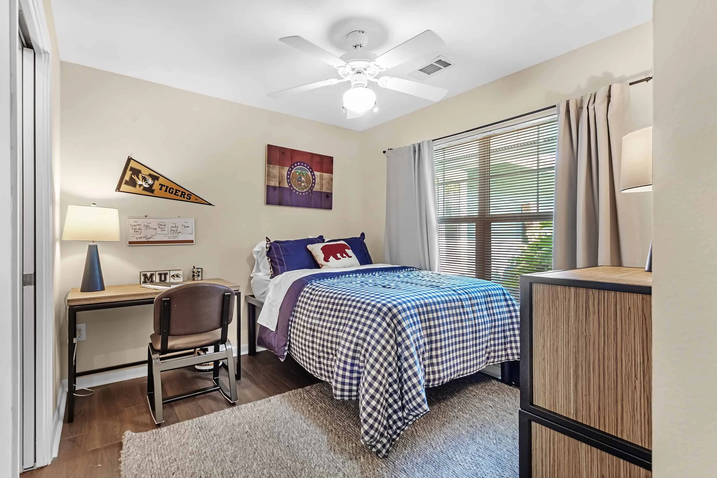 A bedroom with a bed, a desk with a chair, a lamp, wall decorations including a pennant and a flag, a window with blinds and curtains, and a ceiling fan with light.