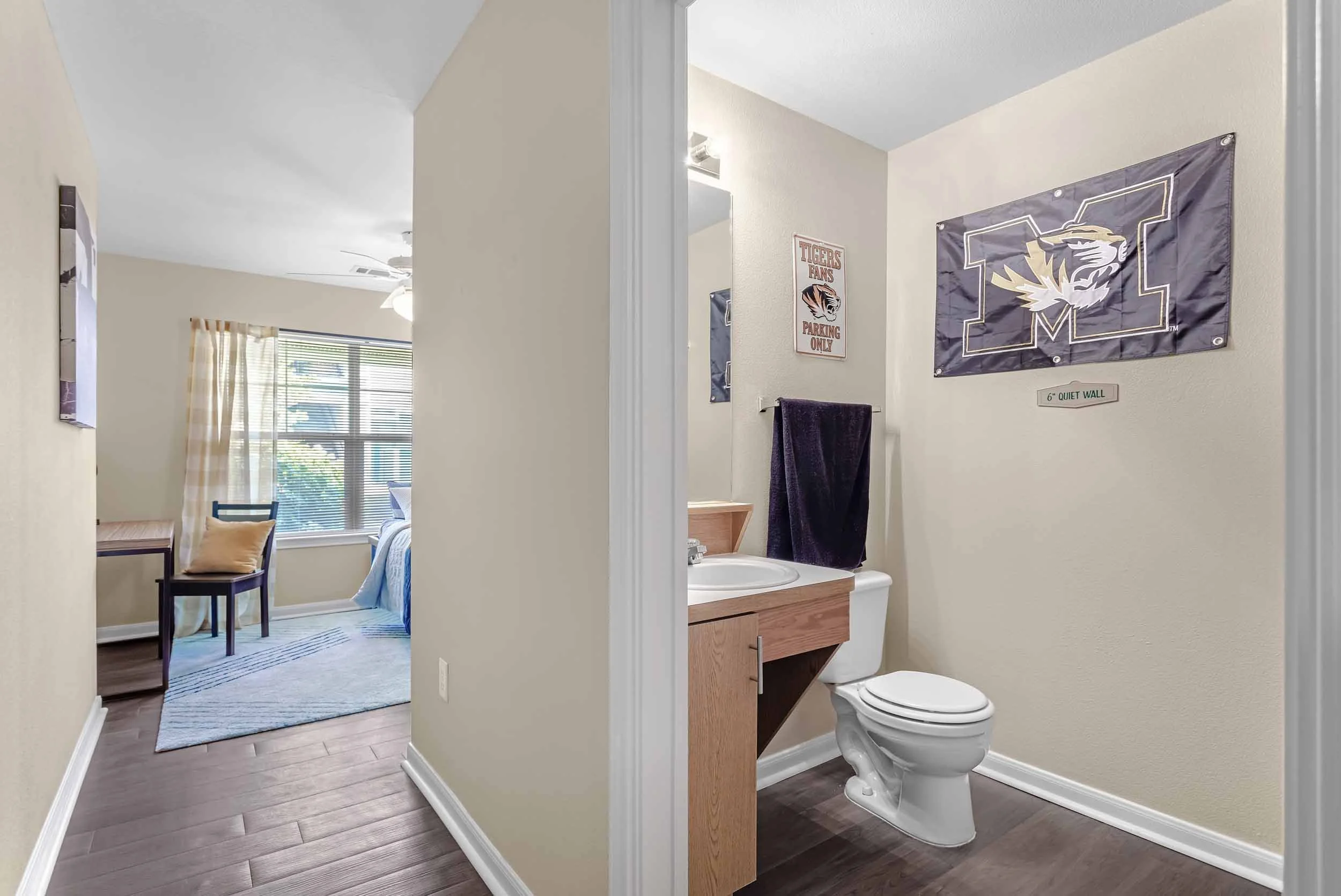 Interior view of a small bathroom with a toilet and wooden cabinet, connected to a bedroom with a chair, window, and bed.