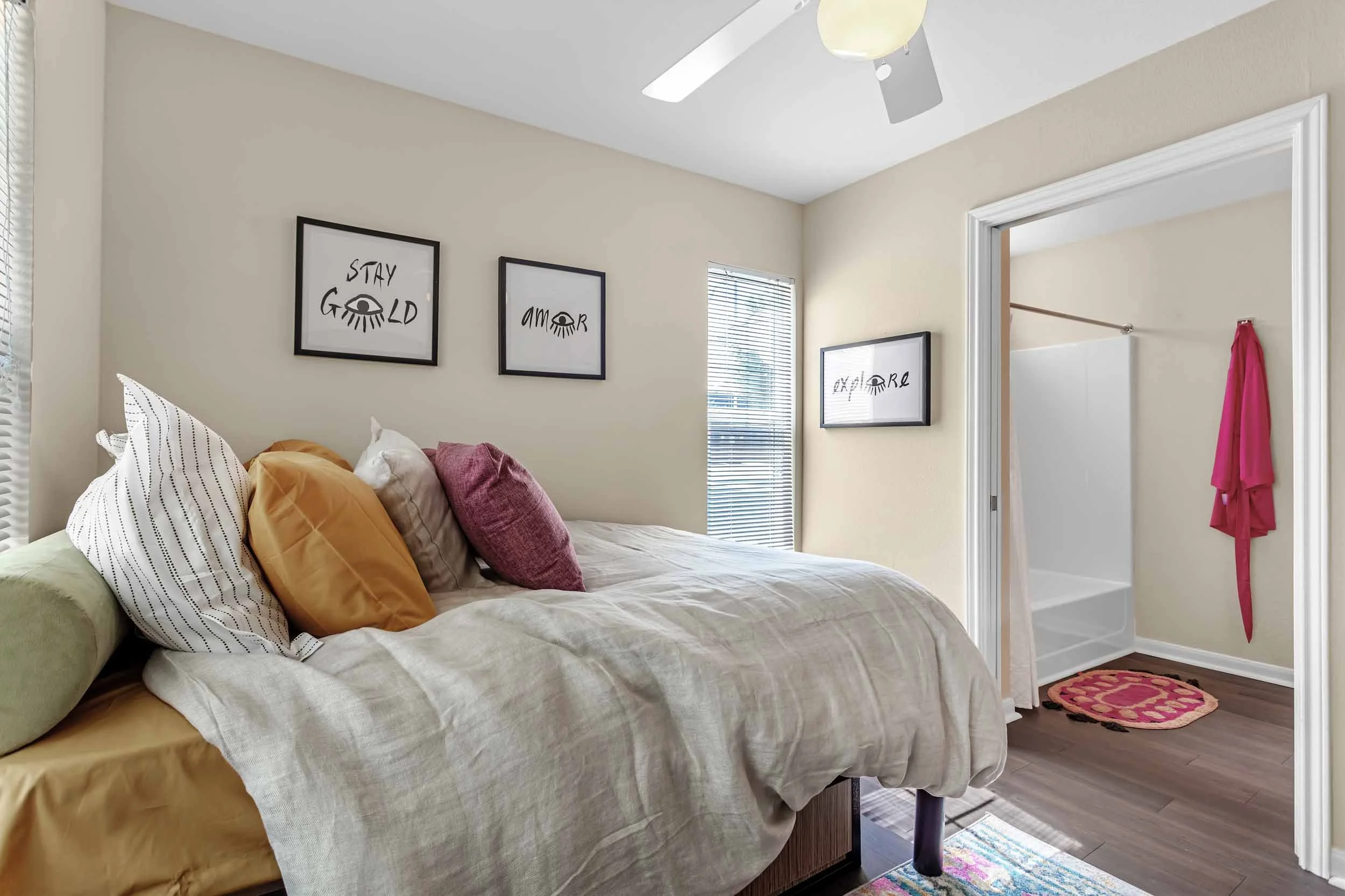 A bedroom with a bed decorated with yellow, white, and pink pillows. Artwork on the wall displays motivational phrases with eye illustrations, and an open doorway reveals a bathroom with a pink towel hanging and a small pink and orange rug on the bat