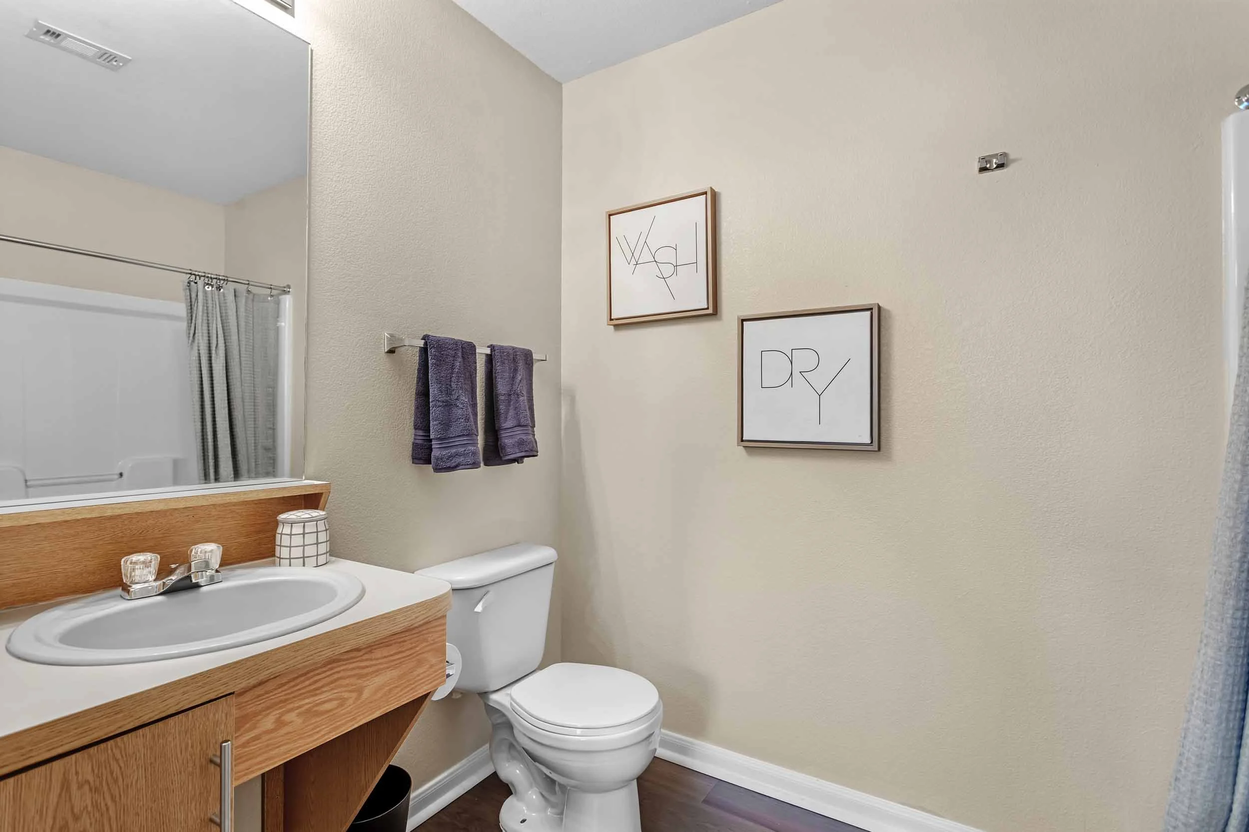 A small bathroom with a white toilet, a wooden vanity with a white sink, gray towels, framed wall art that says "WASH" and "DRY," a mirror, and a shower with a curtain.