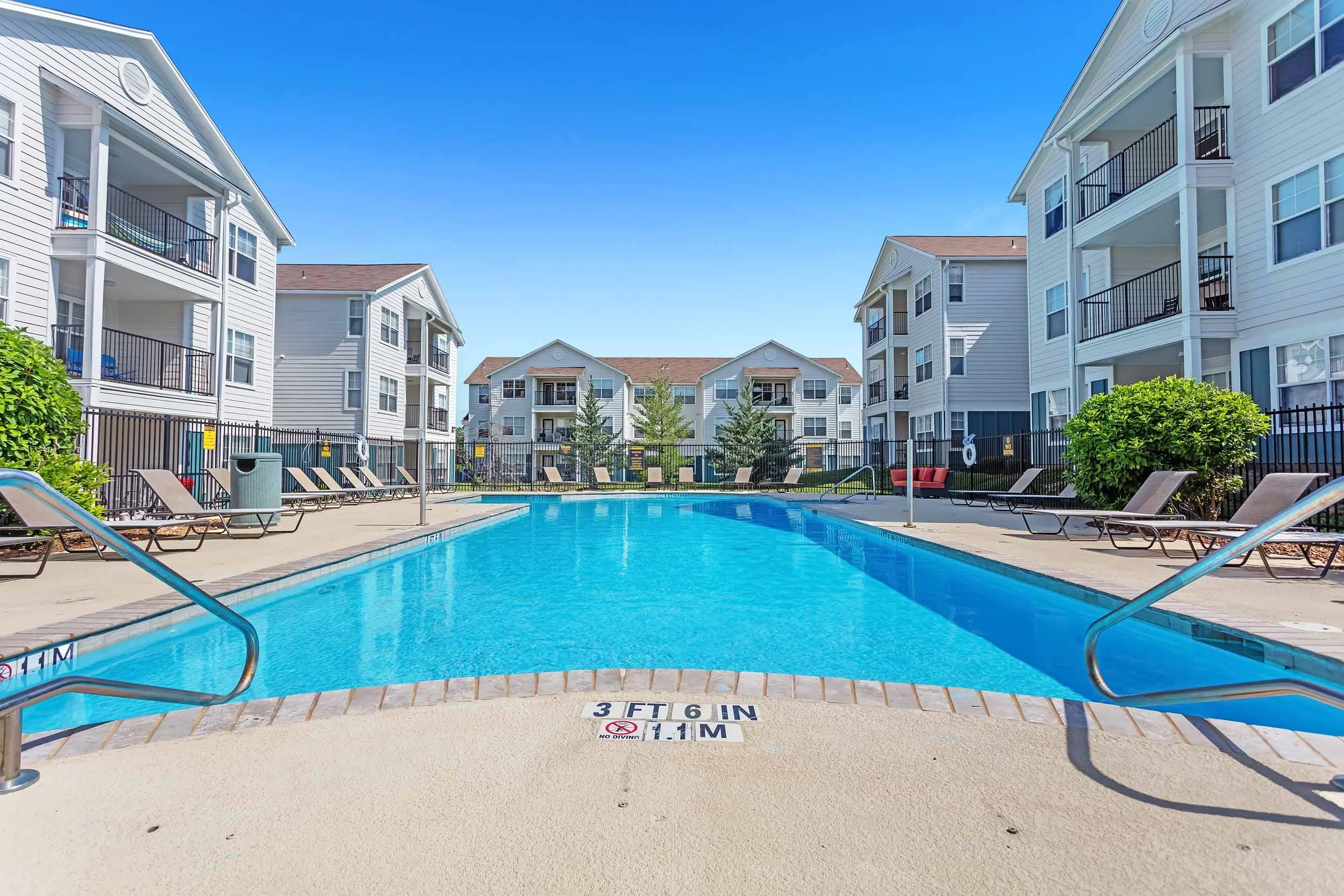 Apartment complex with white buildings surrounding a swimming pool, lounge chairs, and clear blue sky.