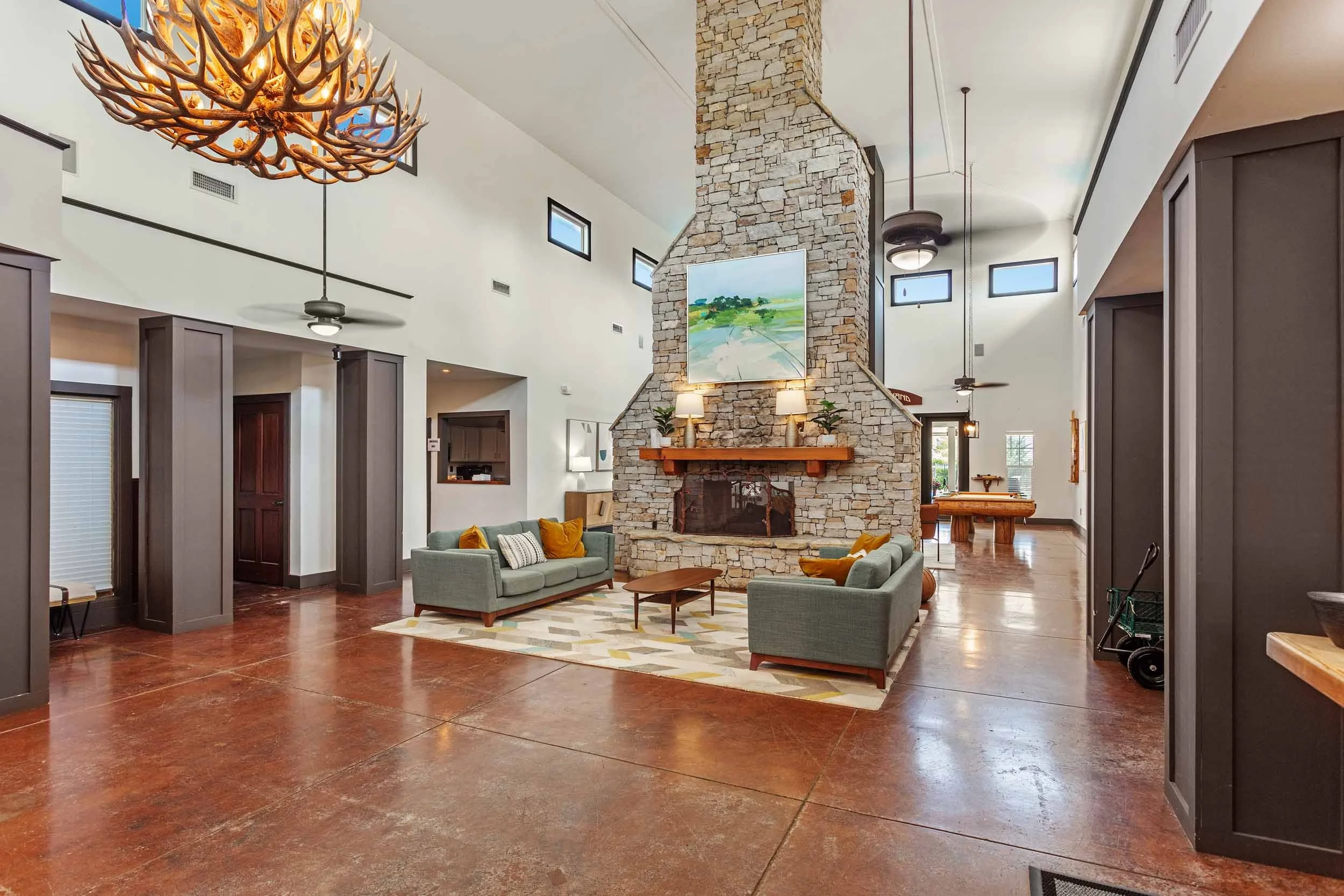 Clubhouse with a stone fireplace, modern sofas with yellow and gray pillows, and a wooden mantel with two lamps and plants. The room has high ceilings, small windows, and open space design.