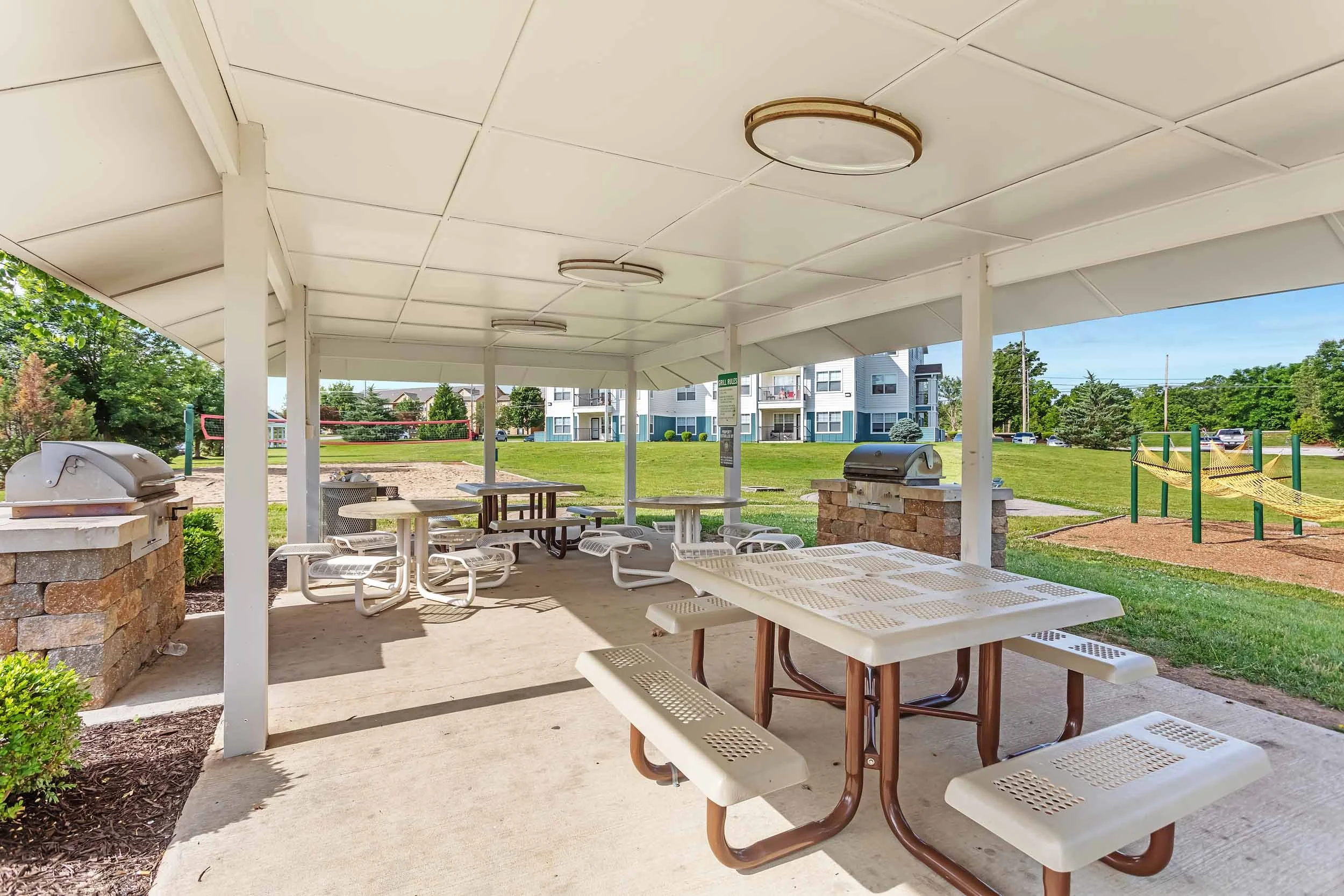 Outdoor covered picnic area with metal tables and benches, surrounded by playground equipment and residential buildings, on a bright sunny day.
