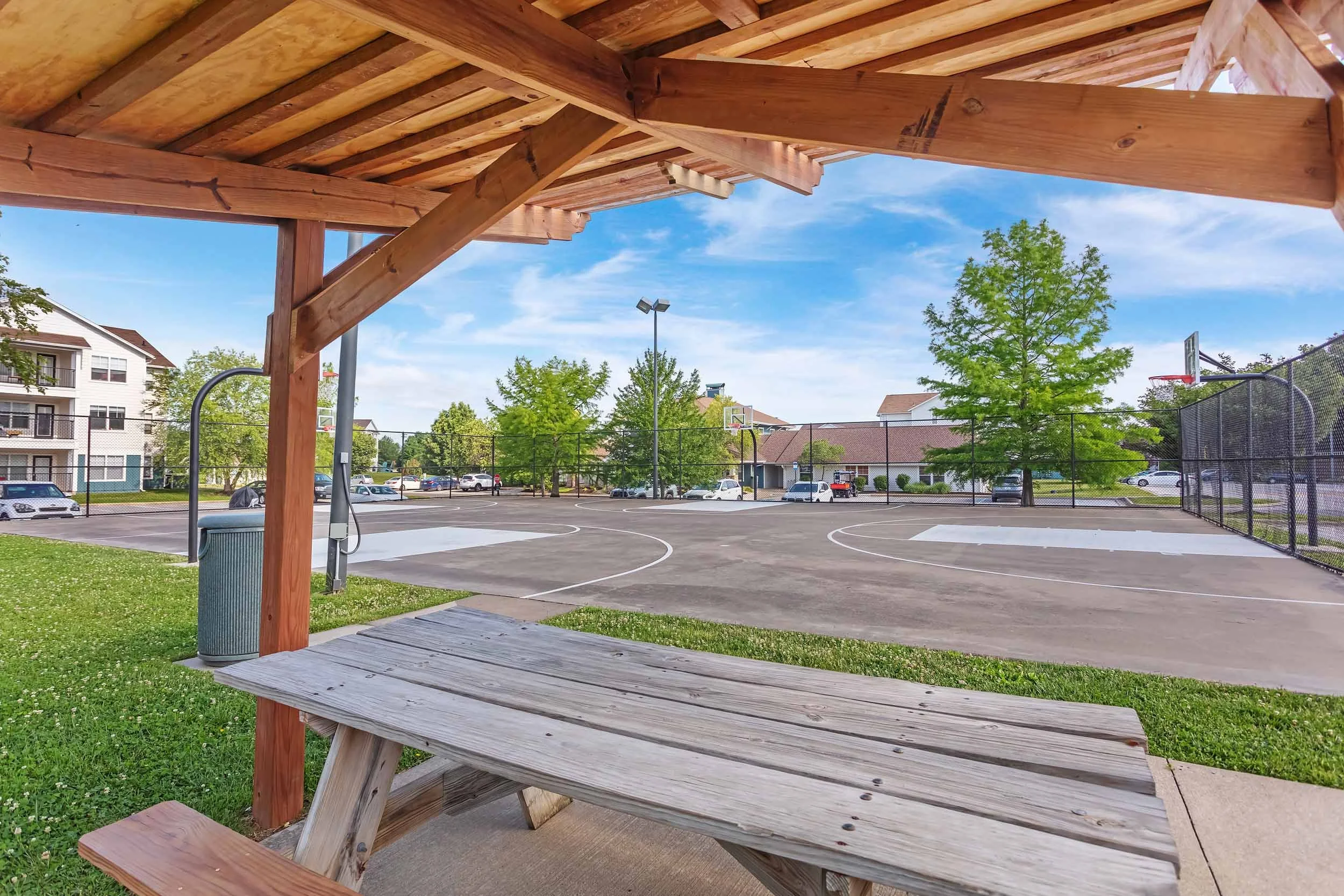 Wooden gazebo overlooking a basketball court with a black chain-link fence, surrounded by trees and residential buildings under a blue sky with scattered clouds.