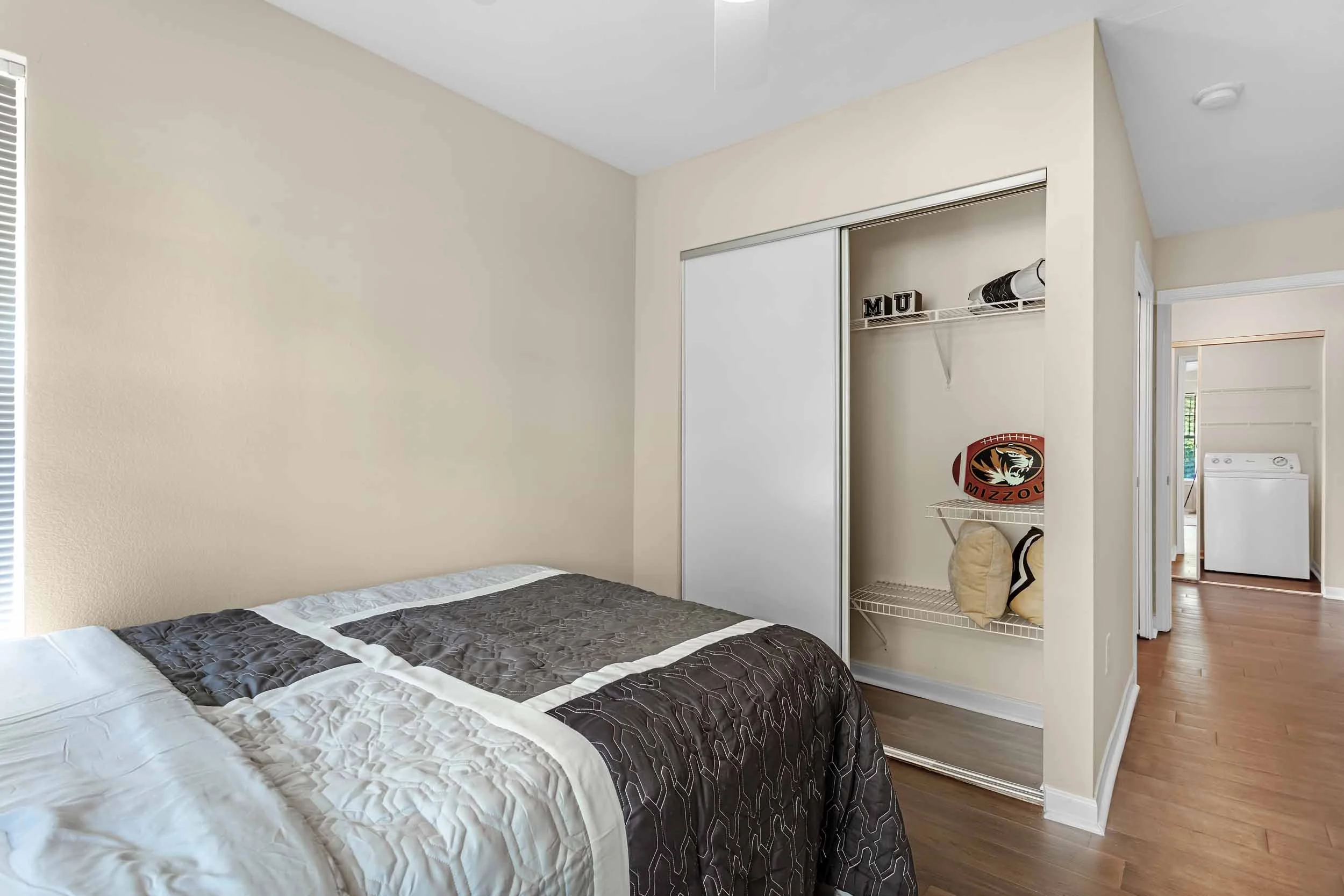 A bedroom with beige walls, a bed with a black and white quilt, and an open closet with a white sliding door. Inside the closet, there are decorative blocks spelling 'MU,' a football with the Missouri Tigers logo, and some bags. To the right, part of